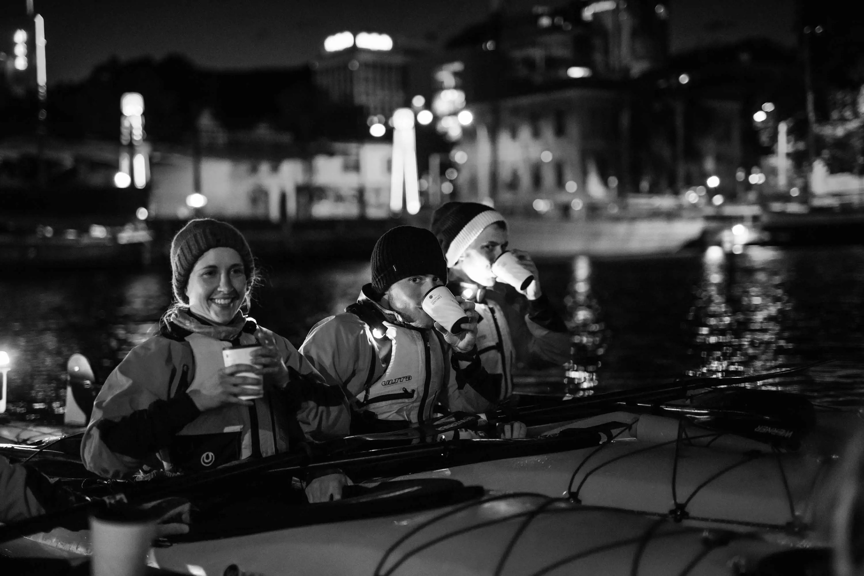 3 people float in small canoes on the water at a port, wearing beanies and sipping at hot drinks in cups.