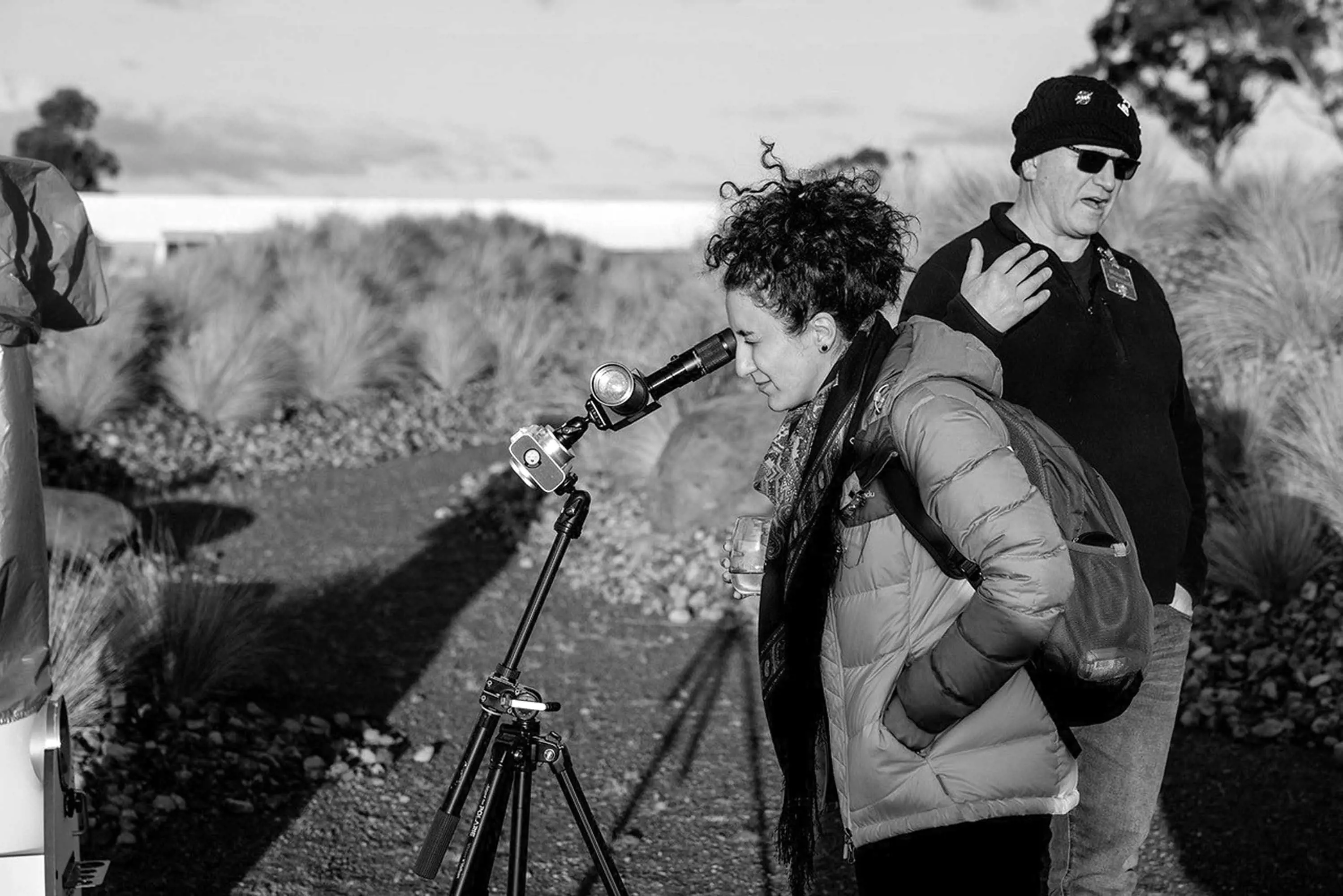 A woman wearing a scarf and a puffer jacket pears into a small telescope set up in a clearing in bushland.