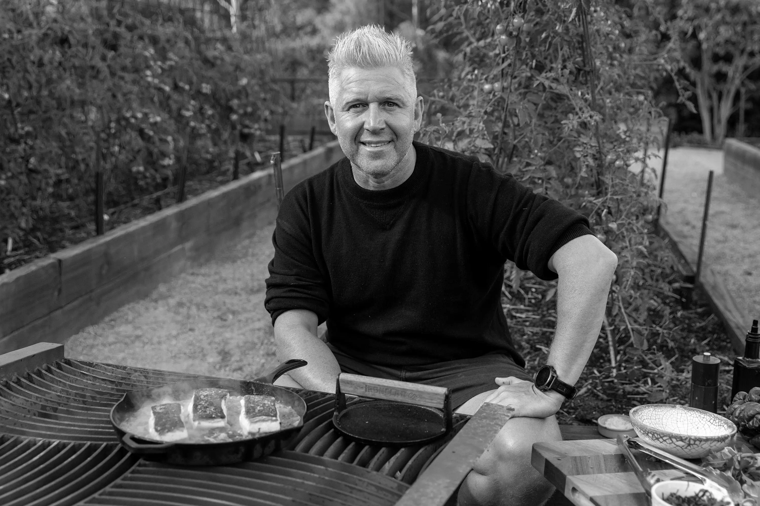 A man in a black shirt sits among raised garden beds, at a barbecue. On the grill is a pan with fillets of fish frying.
