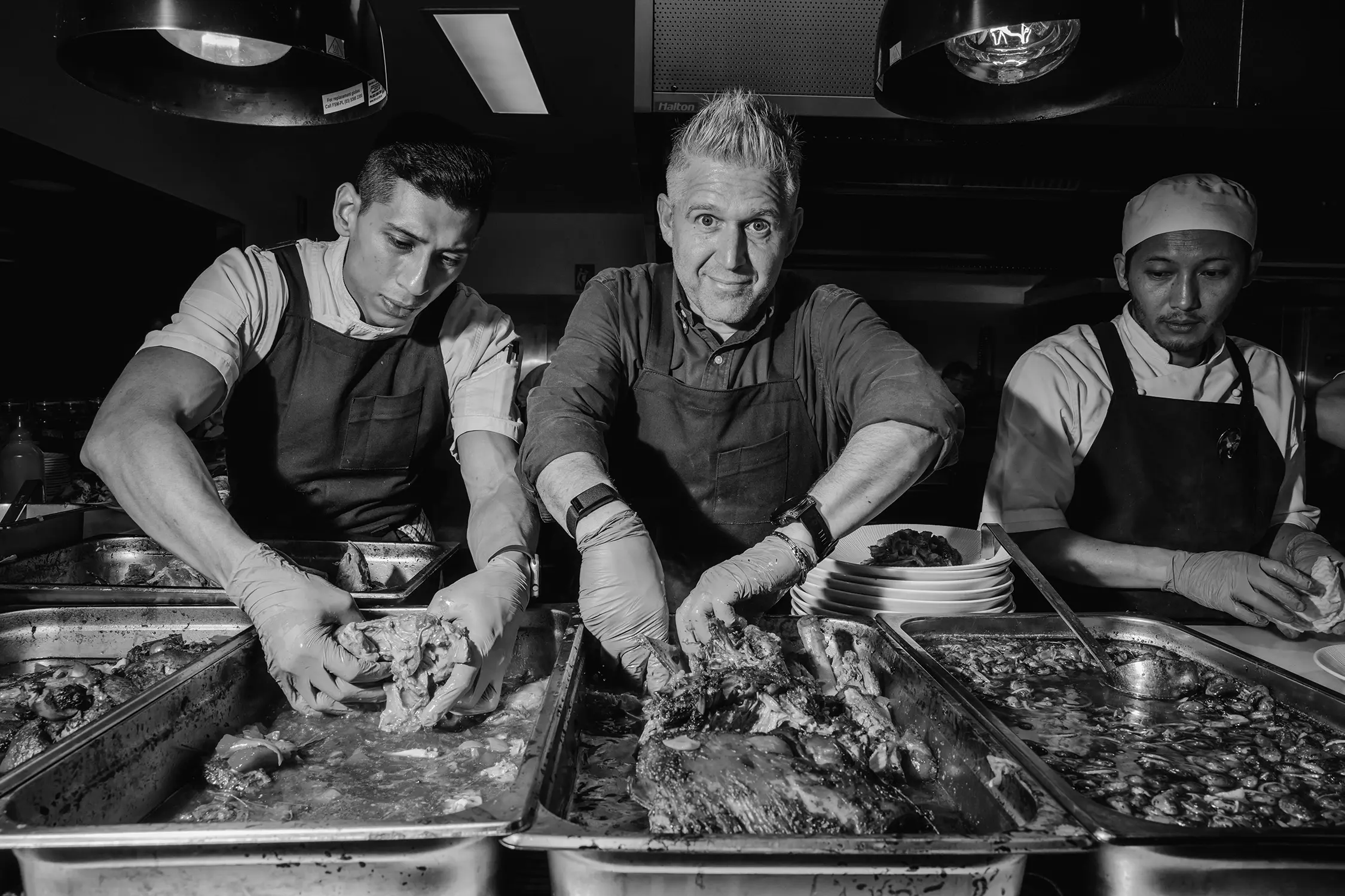 Three chefs wearing aprons stand over a row of huge bain marie trays, filled with delicious fall-apart meat and pan juices.