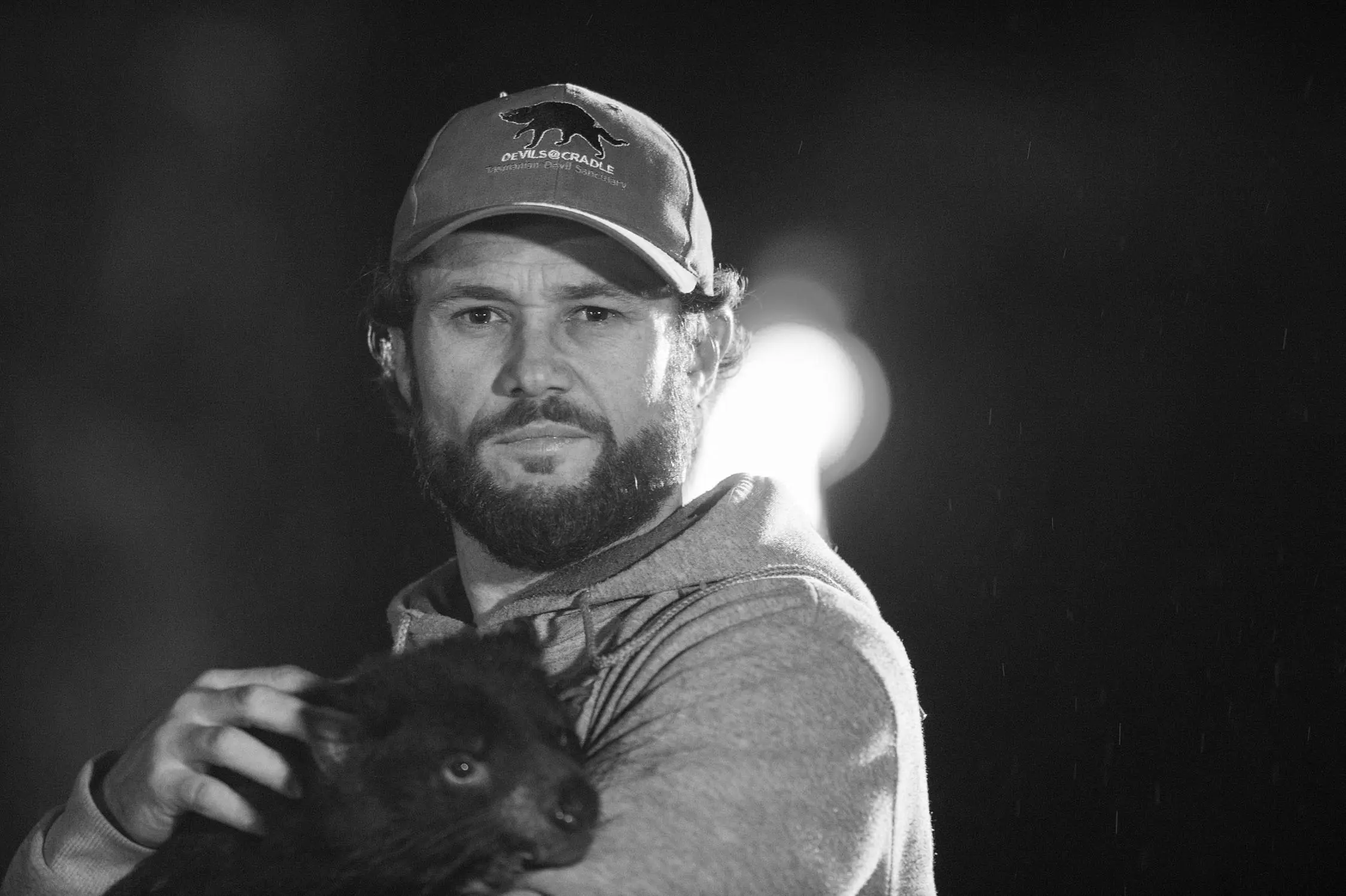 A man in a Devils@Cradle cap holds a baby Tasmanian devil in his arms as he looks into the camera.