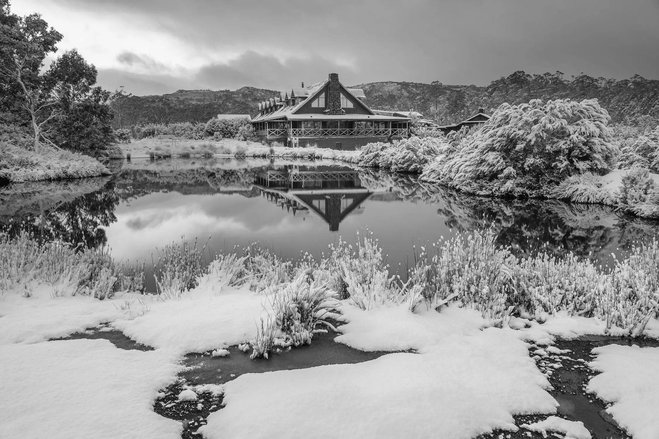 A snow-swept landscape with an alpine lodge in the background. The water is mirror-still, reflecting the scene and the sunset sky above.