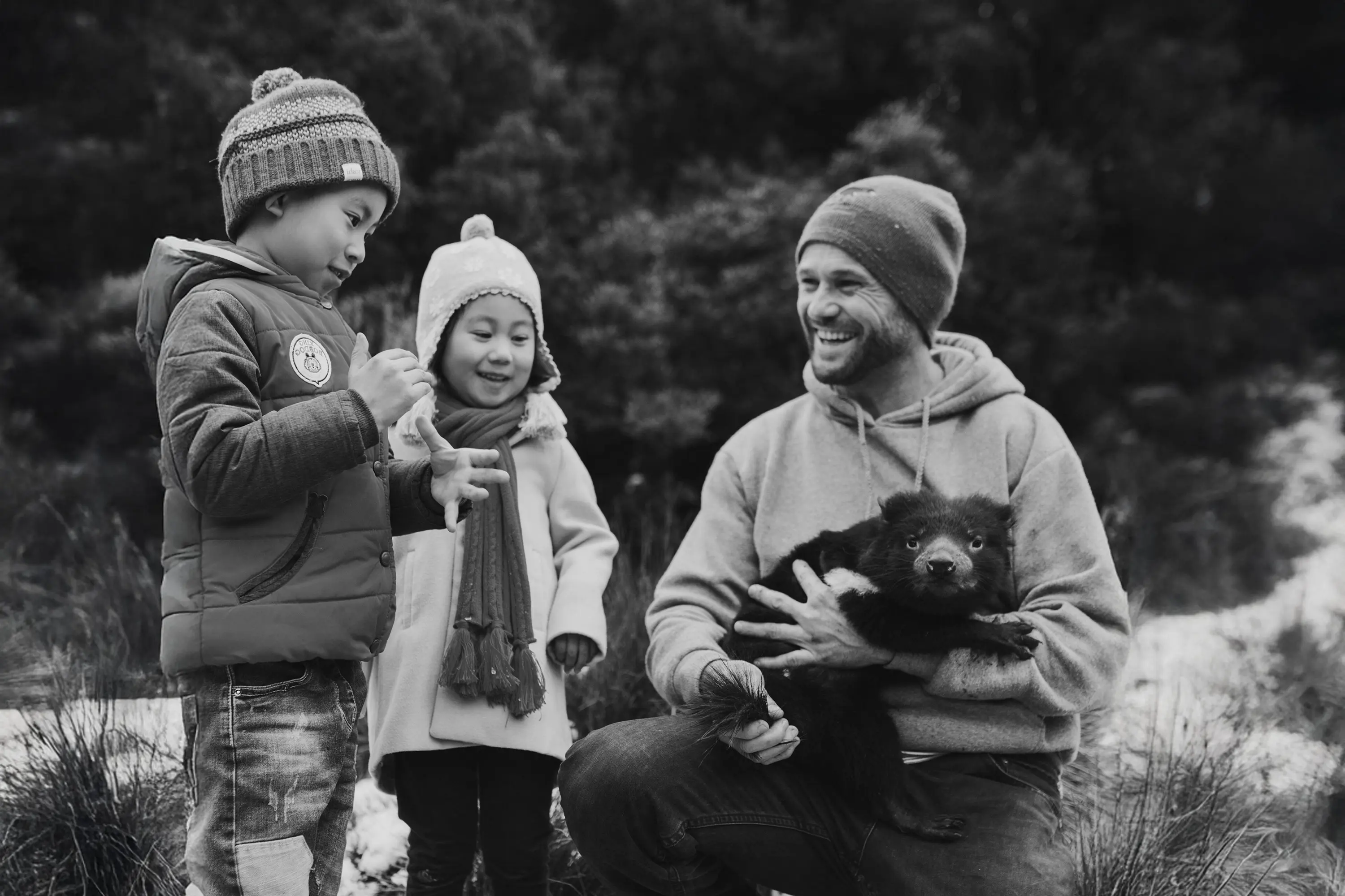 A man in a beanie and hoodie holds a large Tasmanian devil with thick black fur as two small children watch on.