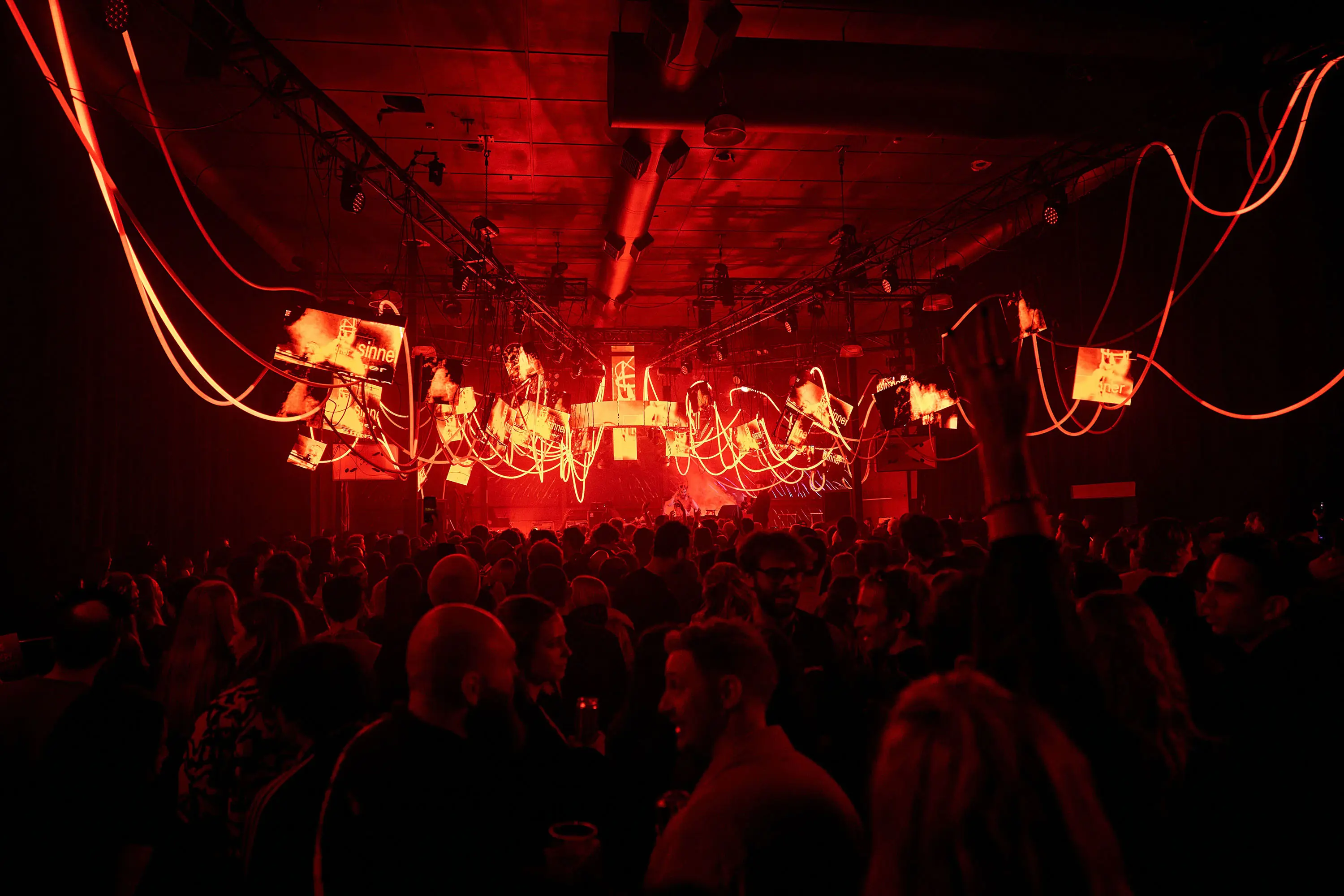 An urban warehouse-style party, bathed in red light. Along the roof are strung glowing ropes and illuminated screens.
