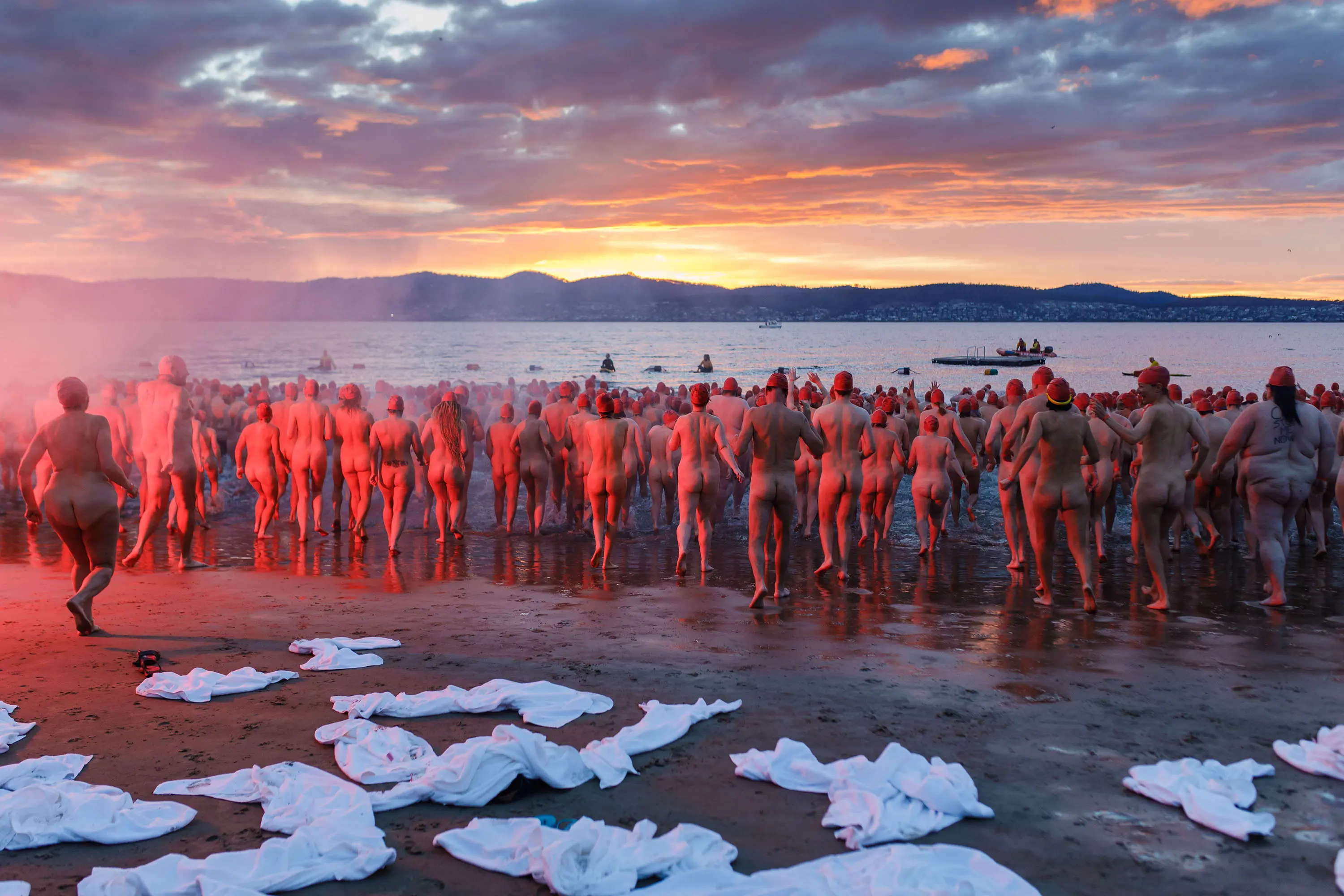 A huge crowd of people, naked except for red swimming caps, run into the water. White towels are strewn on the sand behind them.