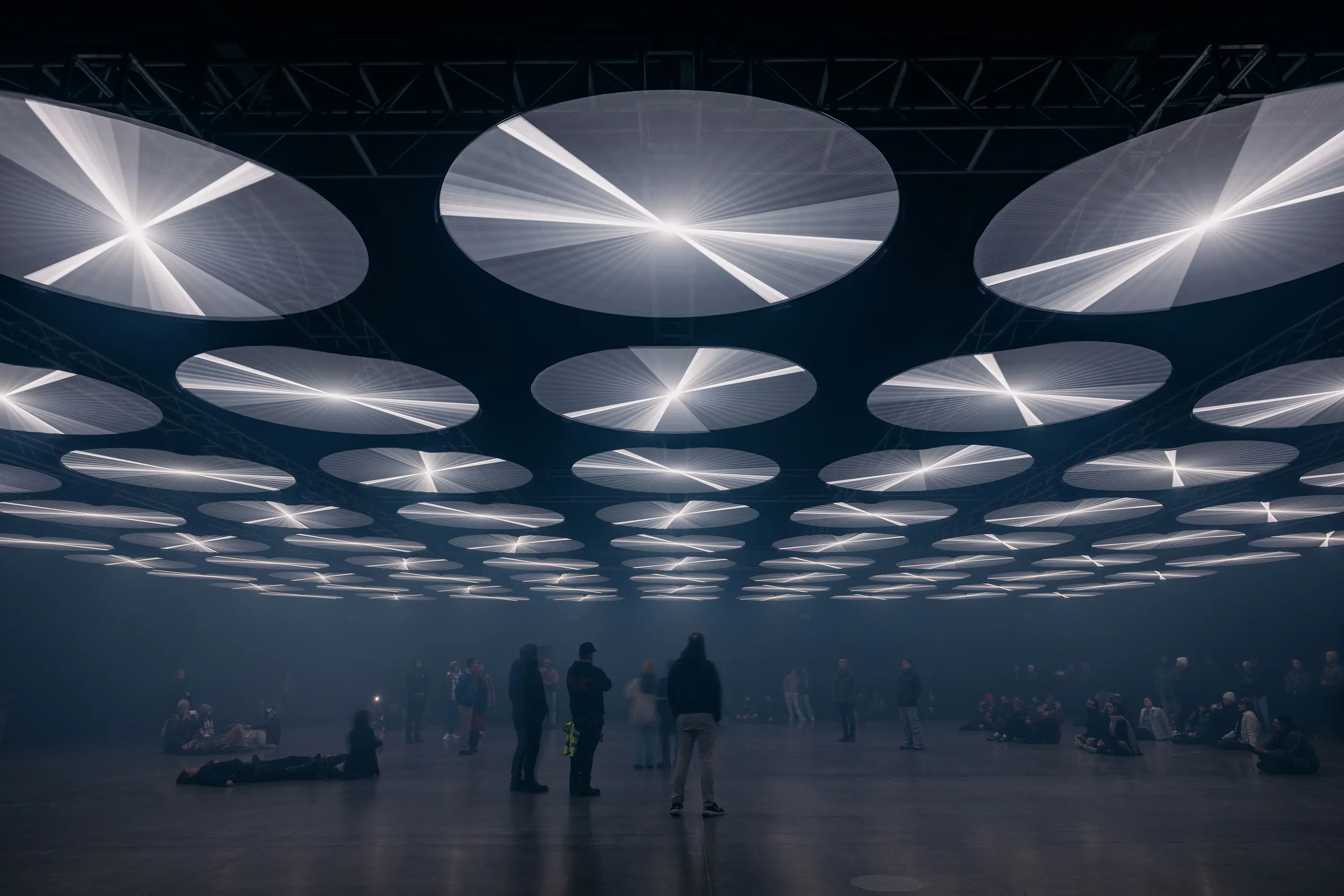 In a huge warehouse space, people are standing, sitting and lying down, gazing at the roof which is covered in circular fan-like light features.