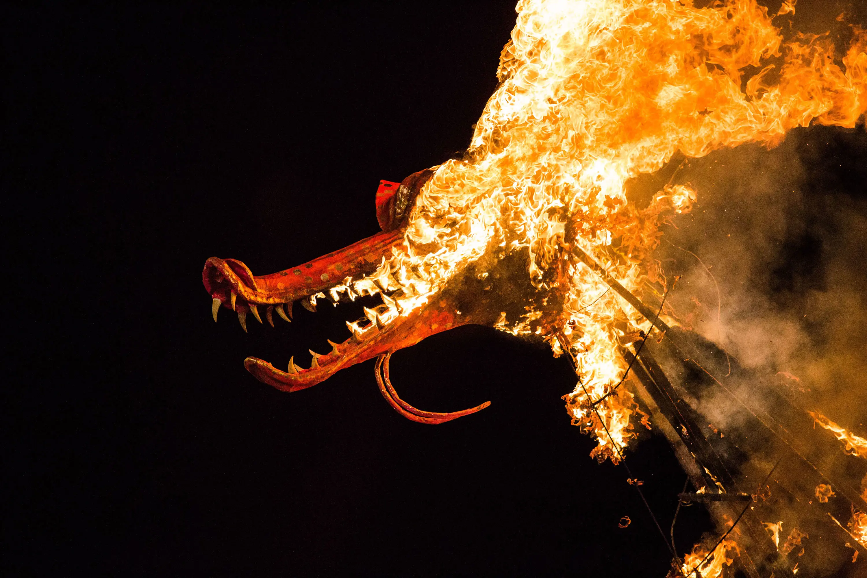 A huge wooden dragon sculpture is on fire, flames pouring from between its toothy jaws.