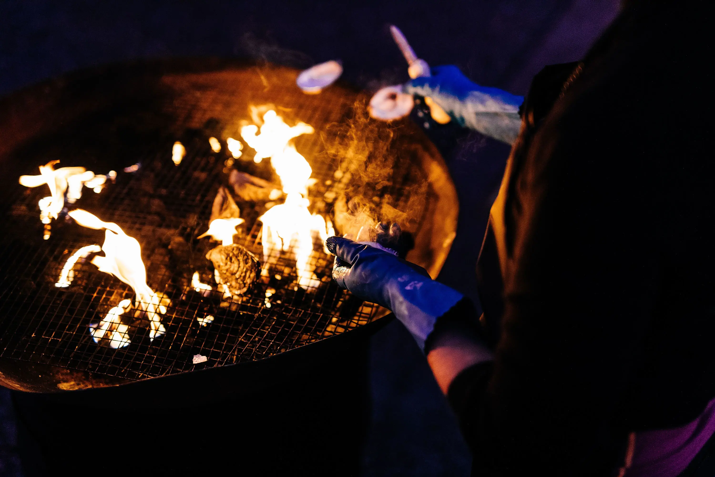 A person barbecues shellfish over an open firepit, covered in a mesh grille.
