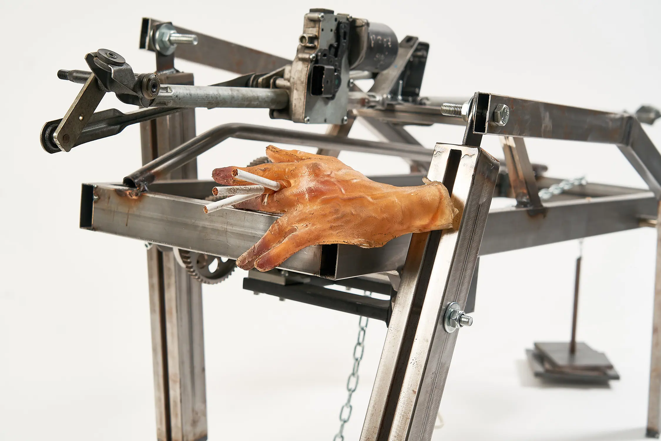 A sculptural robotic metal art piece, featuring pistons, levers, and a disembodied hand holding 3 cigarettes.