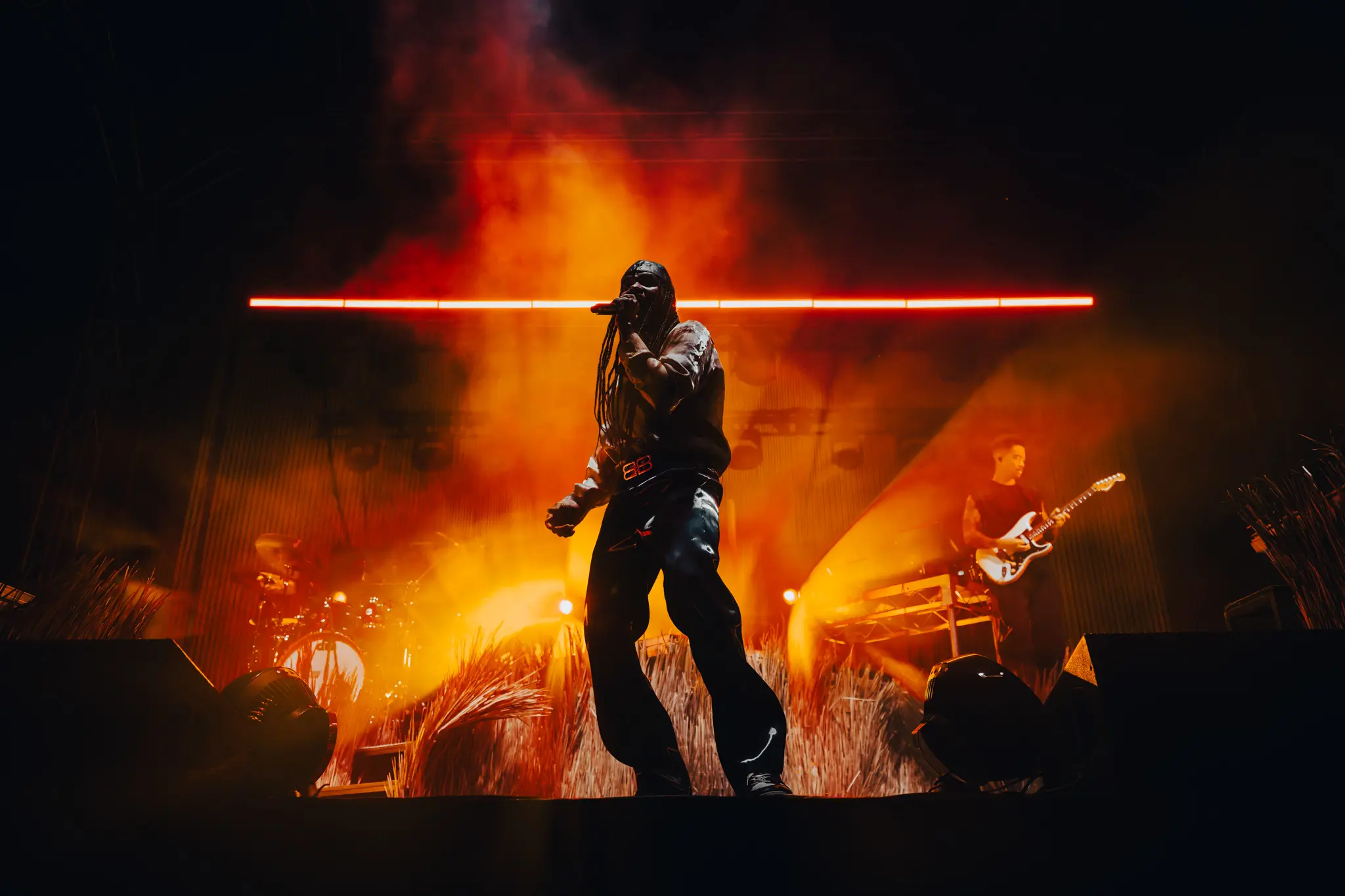 A man stands on a stage, microphone to his mouth, mid-performance. An orange glow of lights is cast onto the stage smoke behind him.