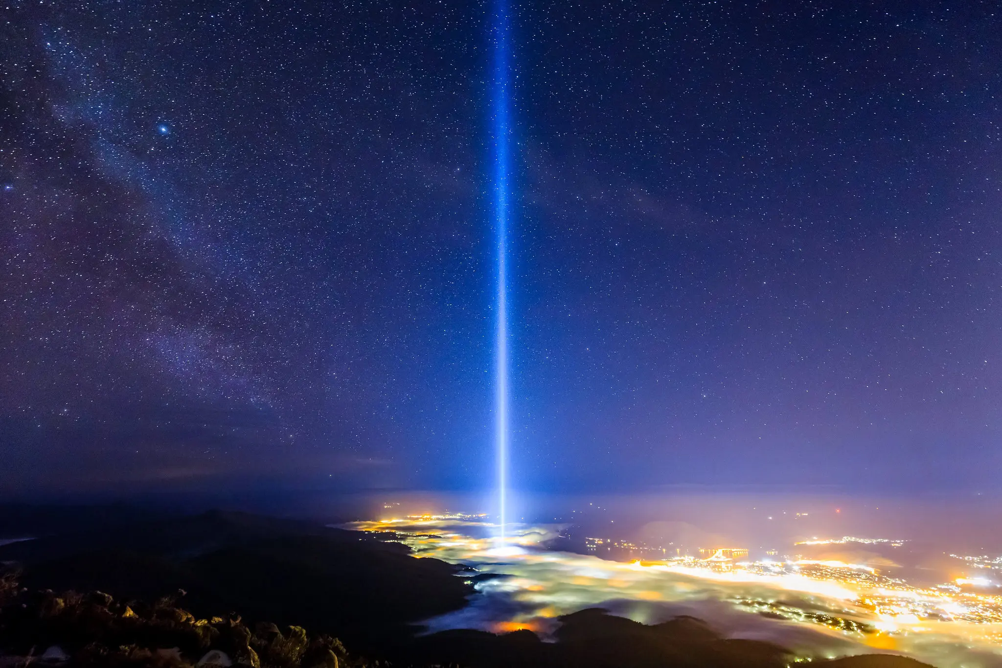 From the top of a mountain overlooking a city, a single strong beam of light is cast into the night sky. The city below is covered in thick fog.