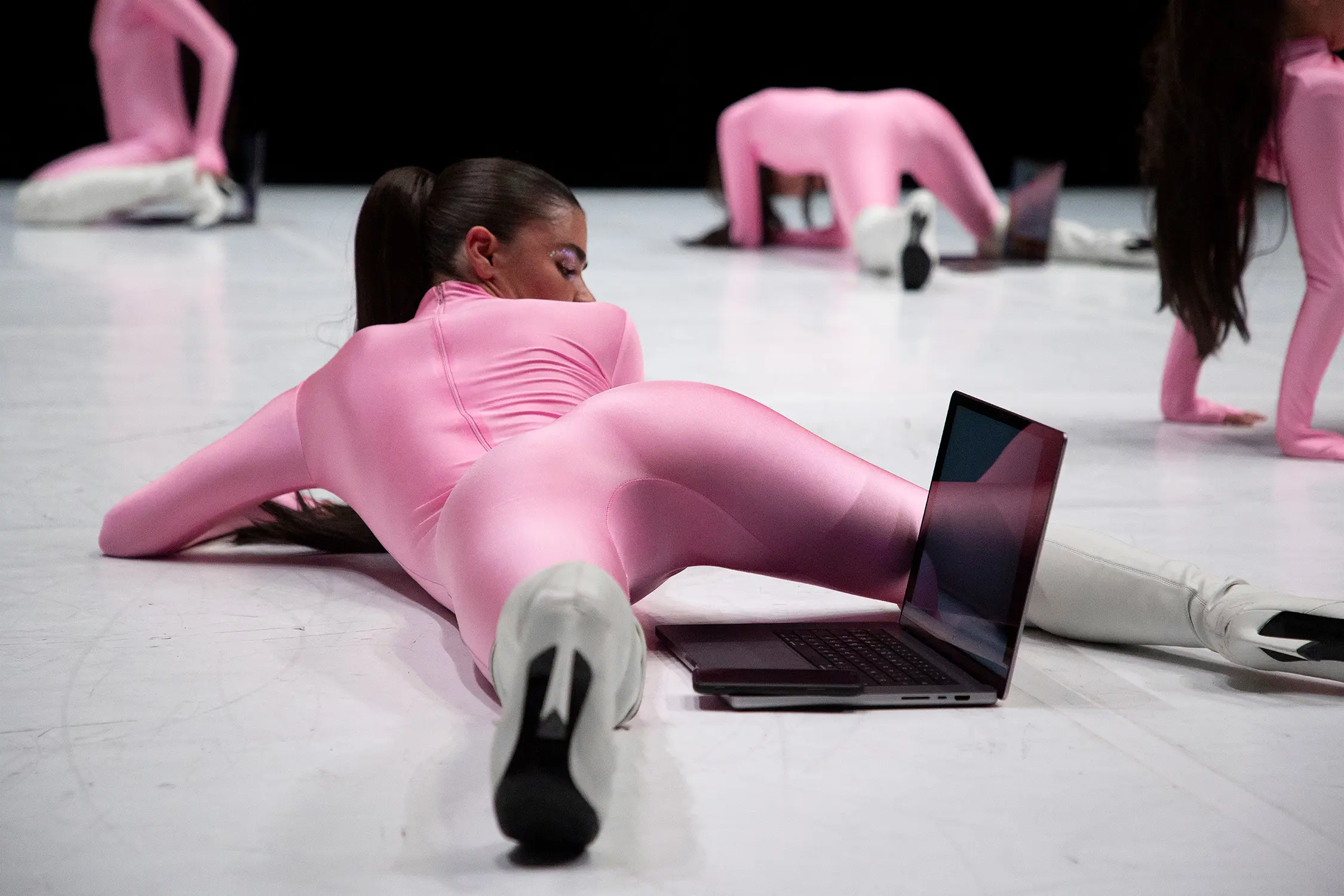 Women in pink skin-tight full-body leotards and knee-high boots writhe around on a stage. One woman has a laptop positioned between her legs.
