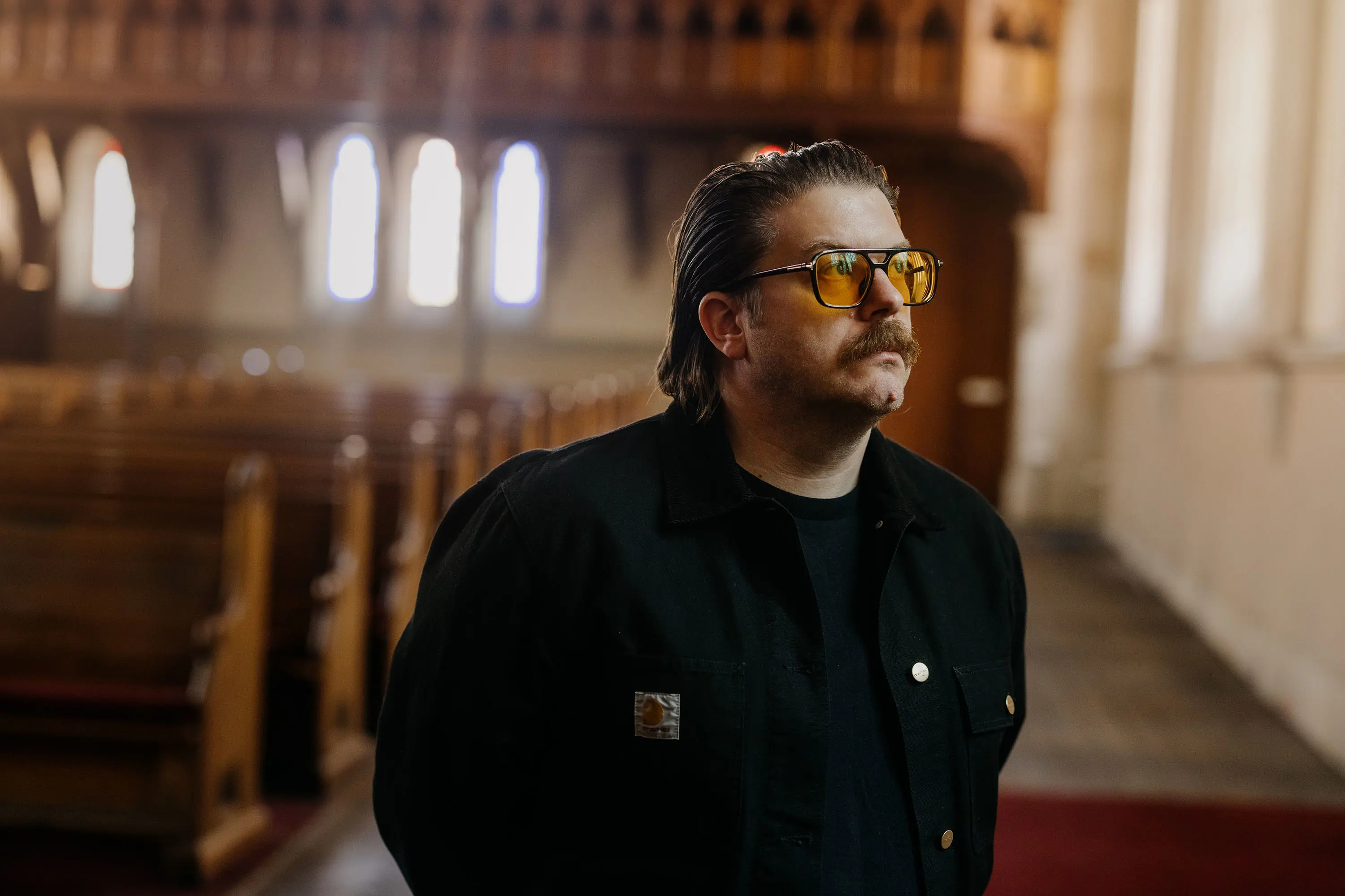 A man wearing a black jacket and large orange-tinted glasses with a moustache and slicked-back hair stands gazing out a window. Behind him, the out-of-focus background of a church with pews and sculptural windows is visible.