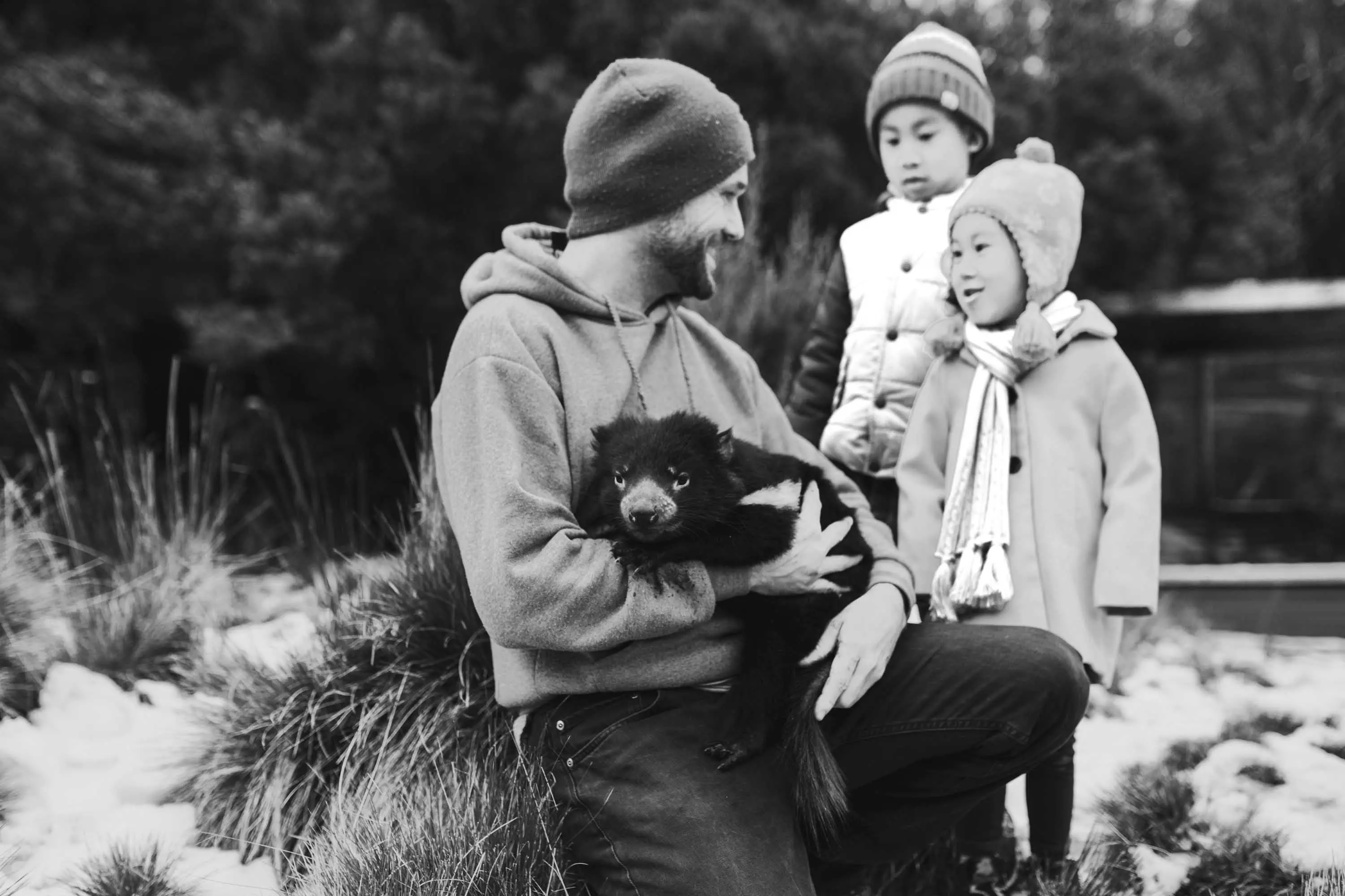 A man in a beanie and hoodie holds a large Tasmanian Devil with thick black fur as two small children watch on.