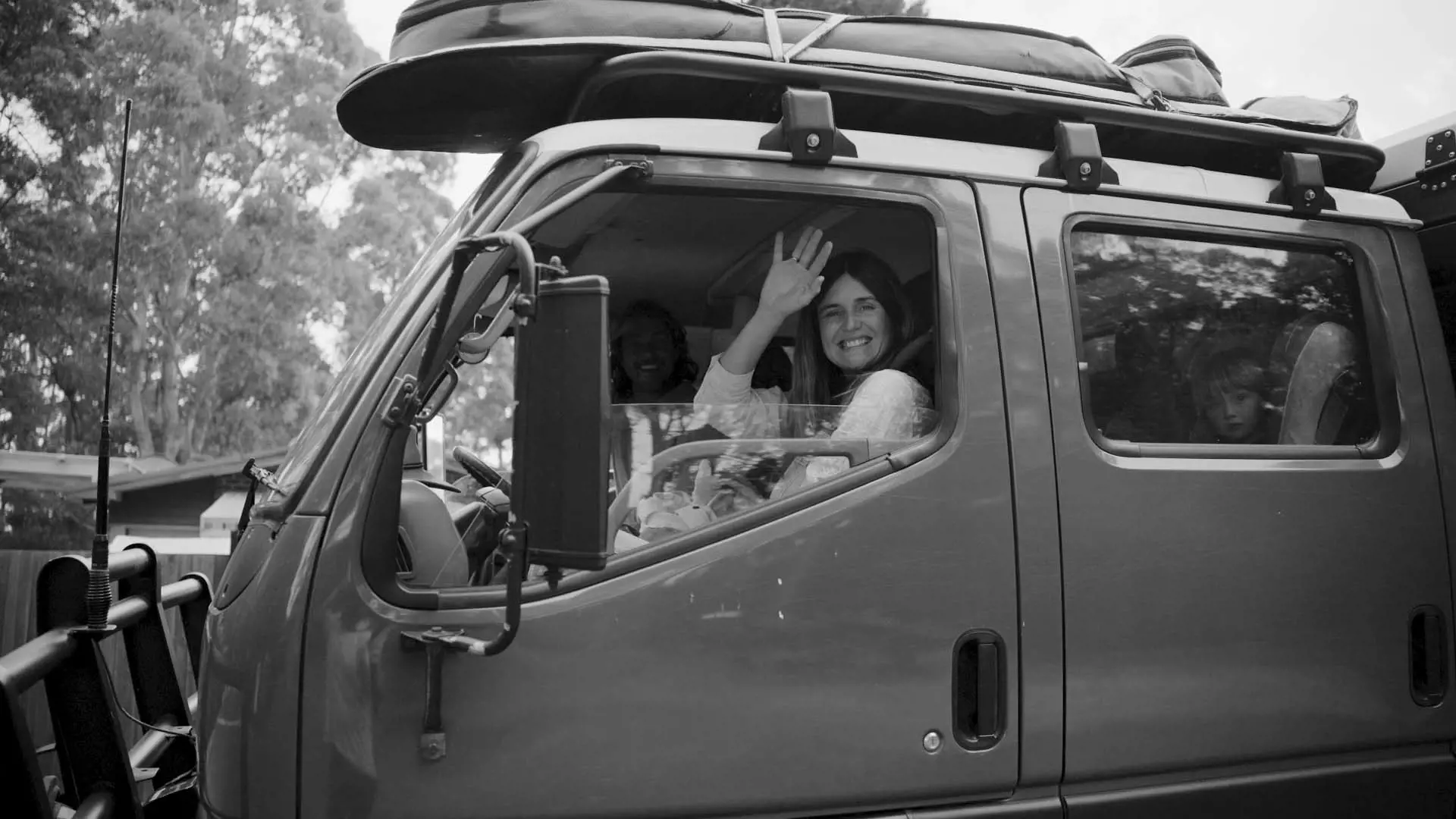 Inside a vehicle, a woman smiles and waves from the driver's seat while a child is seen looking out the window in the background.