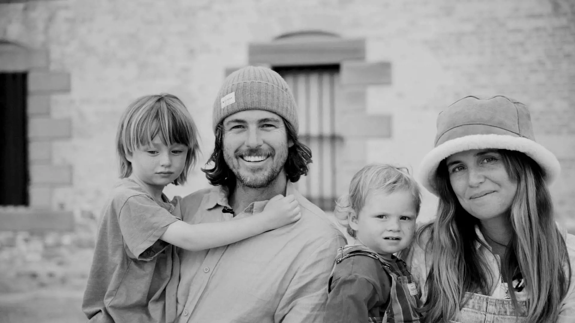 A smiling woman and man stand with two children, one on his hip, in front of a rustic brick building. The group seemingly enjoys a moment together.