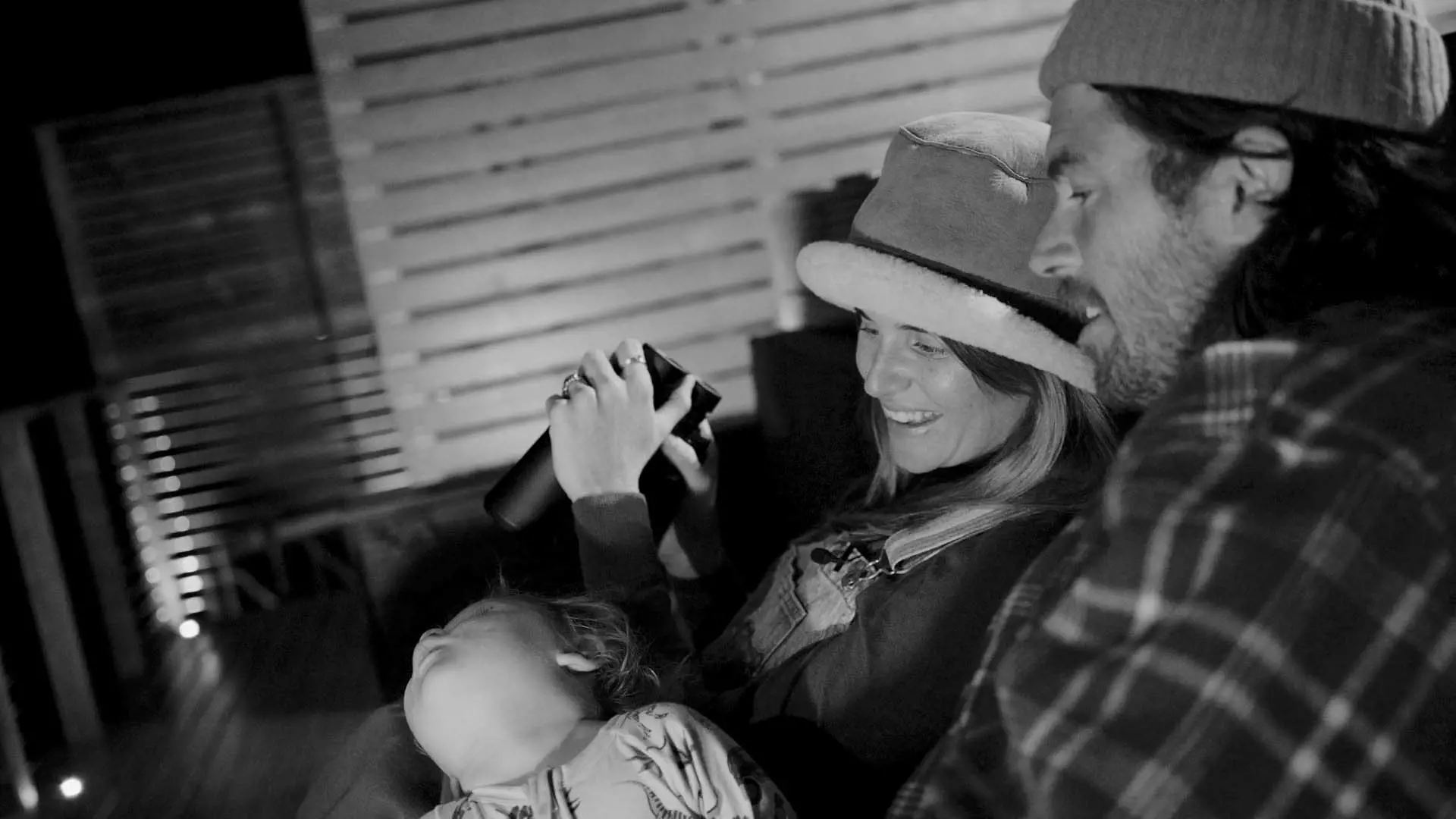 Two adults and a child are sitting together, with one adult smiling and holding up binoculars. The child rests beside them. The setting appears cozy and intimate.