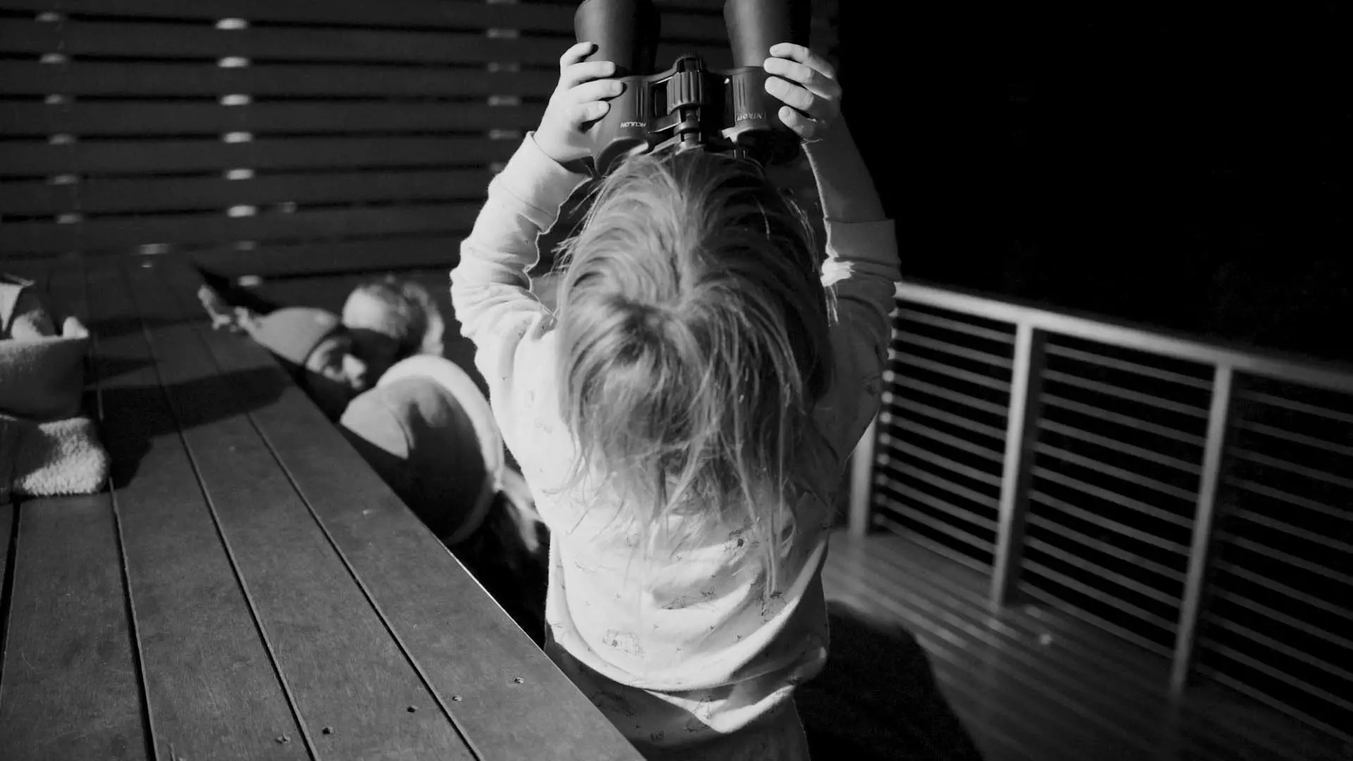 A child wearing pajamas stands on a deck, holding binoculars upside down. The child's long hair falls down their back, and a table is visible in the foreground.