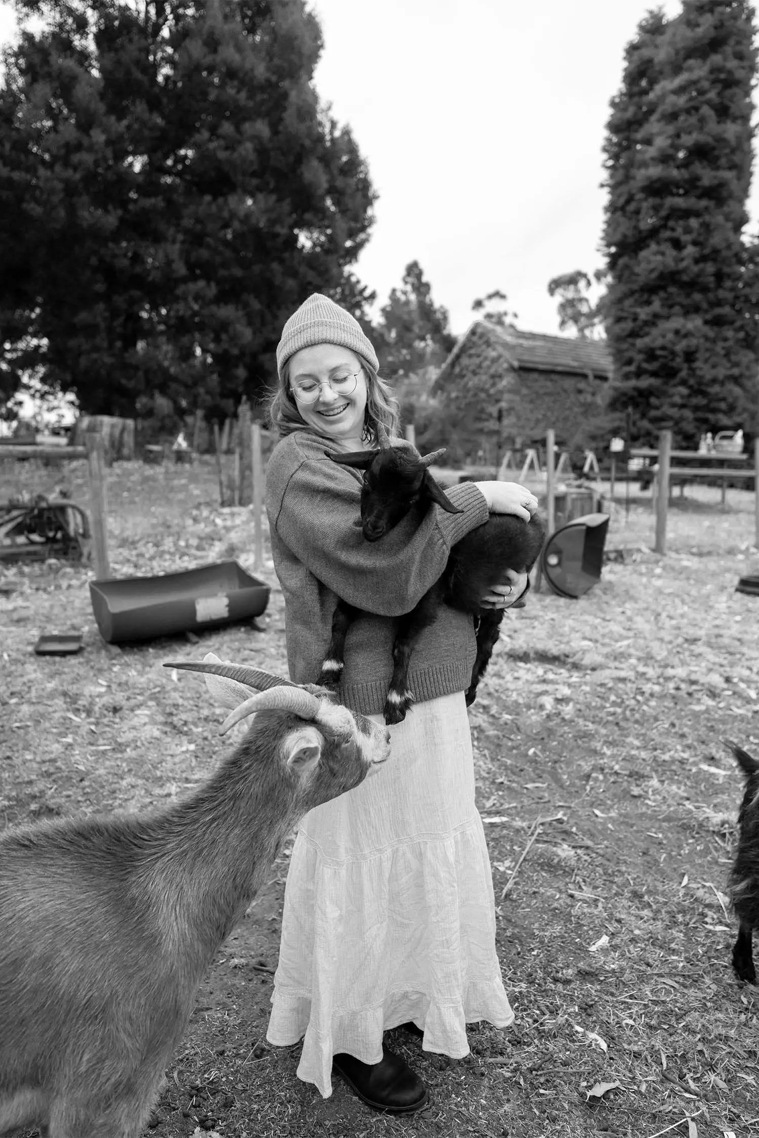 A woman in a sweater smiles while holding a black goat in her arms, as another goat approaches her. The image conveys warmth and connection, and is in black and white.