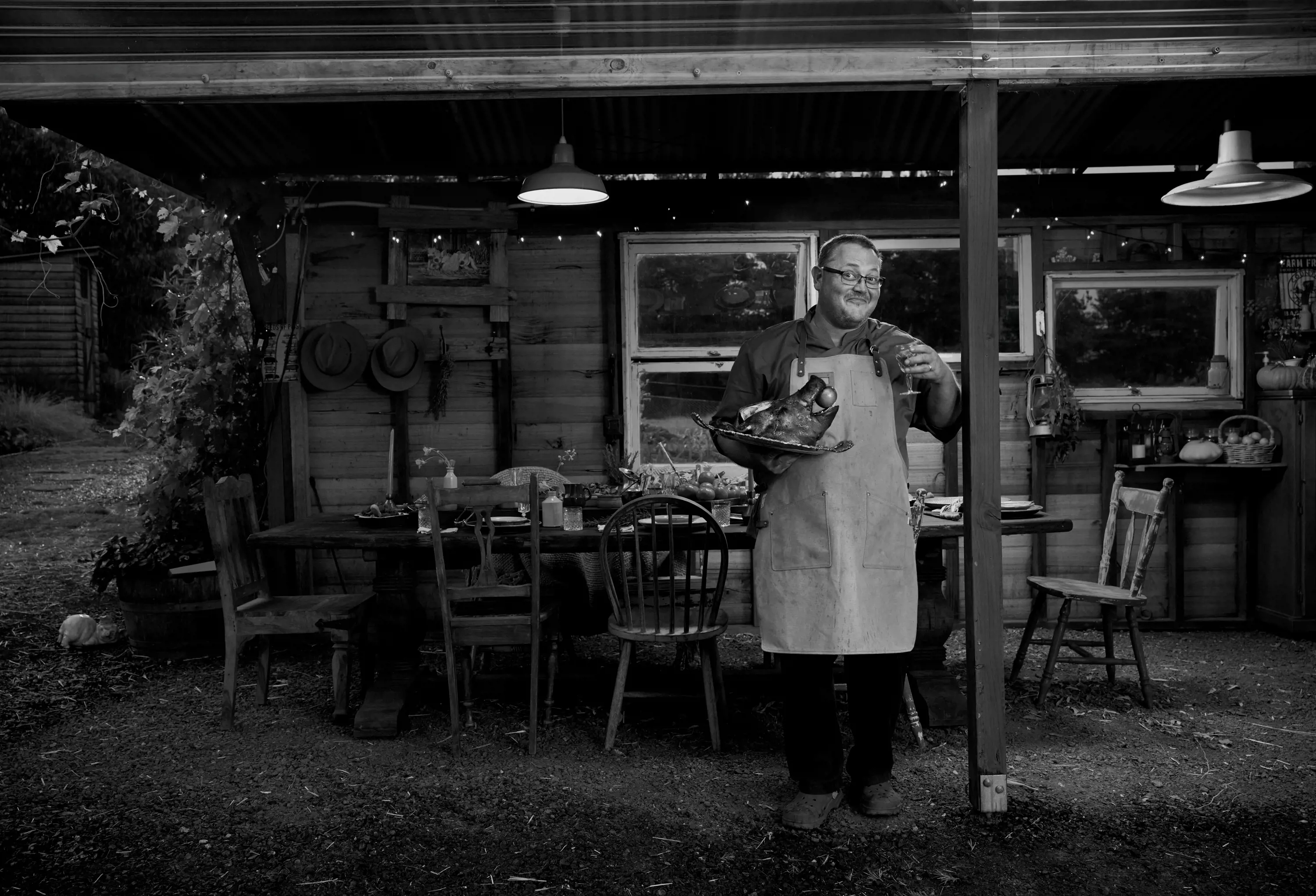 A man wearing glasses and a cooking apron stands under a verandah holding a glass of wine and pig's head on a plate.
