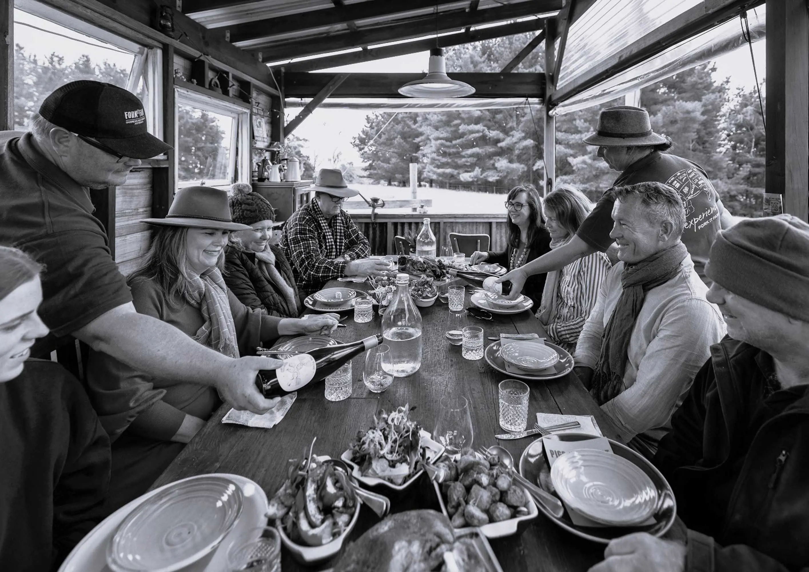 A large group of people sit at a long wooden under a shelter table and enjoy a meal together. 