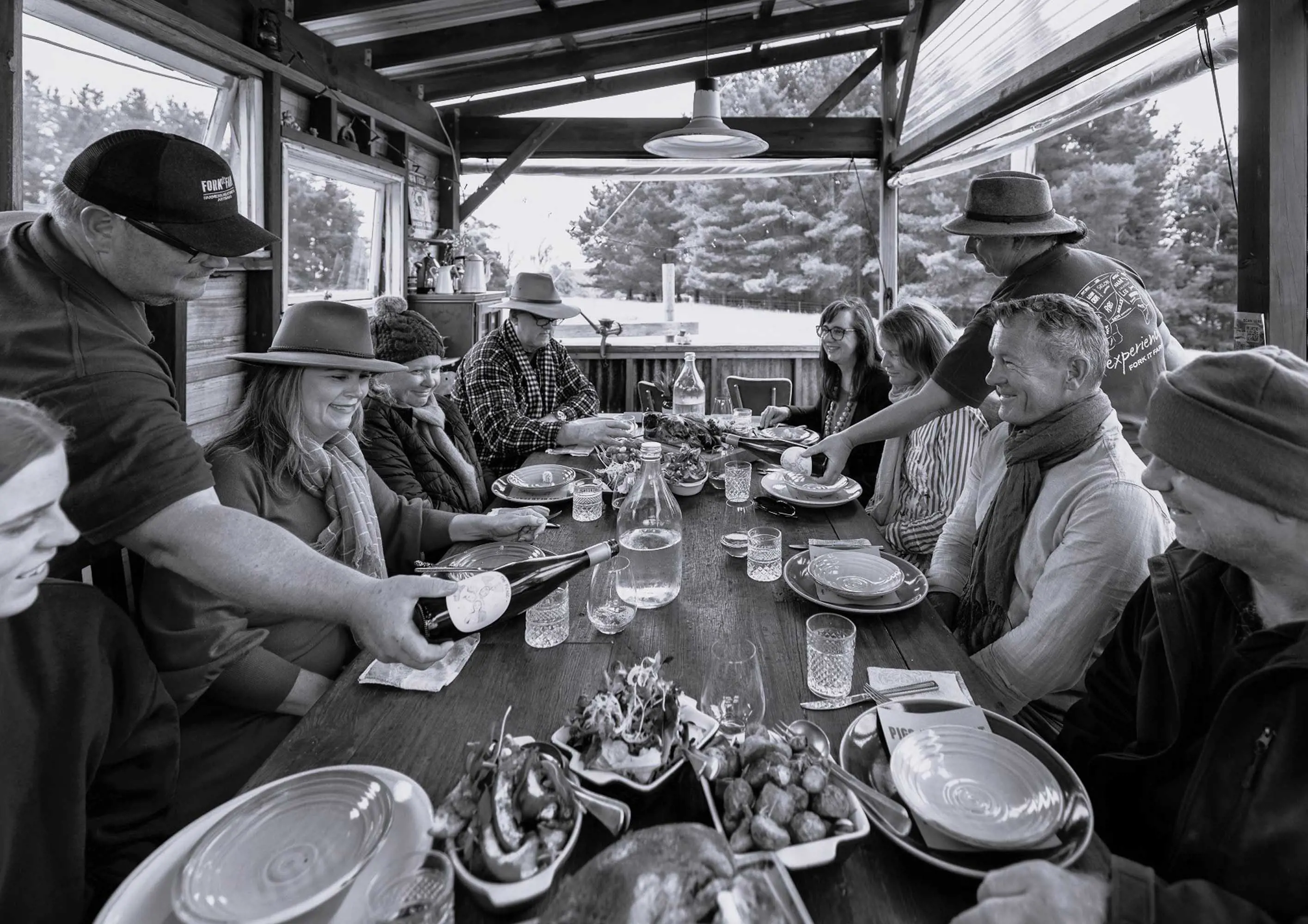 A large group of people sit at a long wooden under a shelter table and enjoy a meal together.