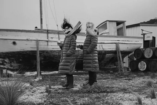 Two people wearing body-length puffer jackets stand in front of a large wooden boat up on stilts near water.