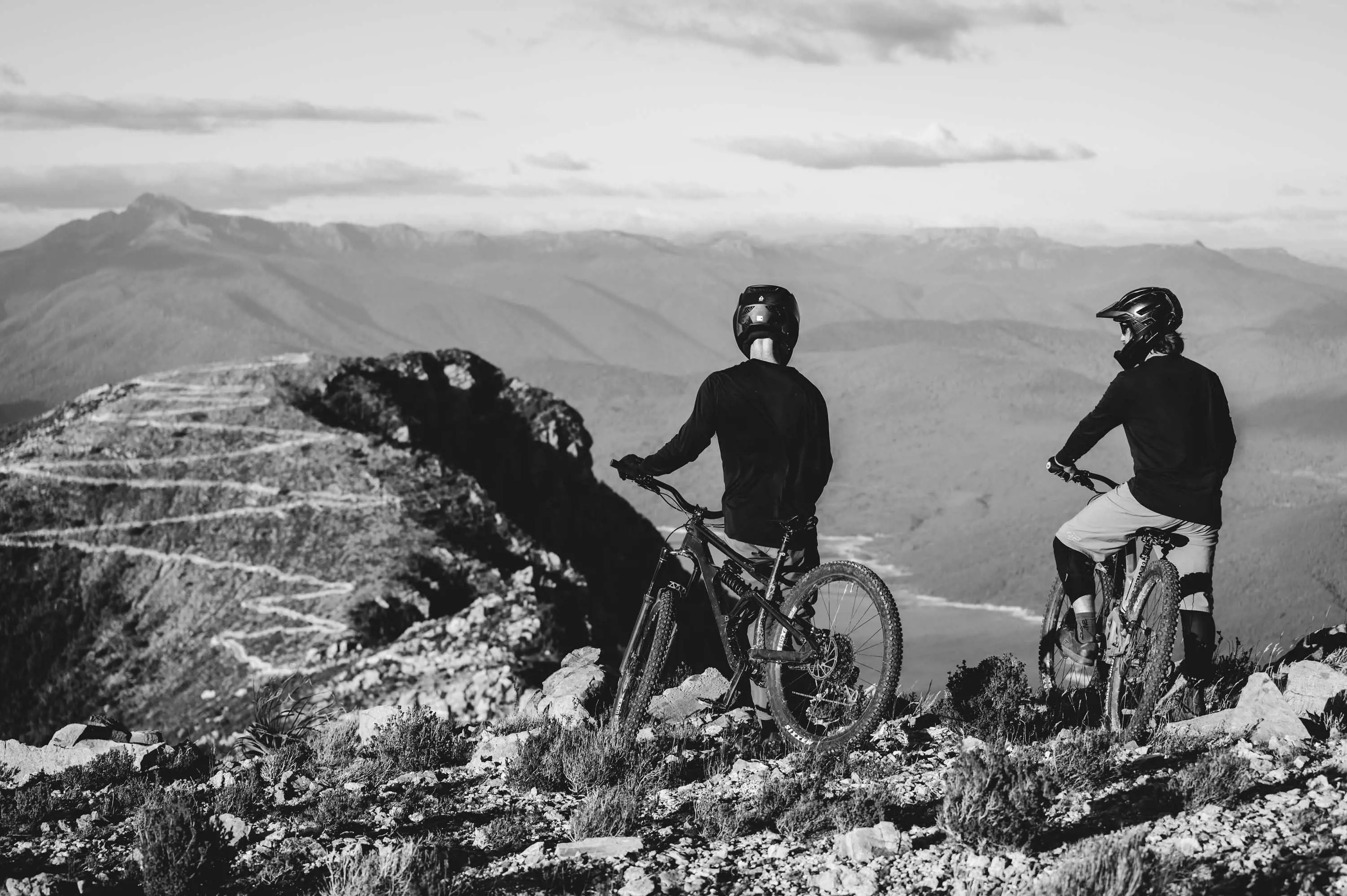 Two mountain bike riders wearing helmets and protective equipment stop to look at the gravel track zig-zagging down a mountain in the distance.