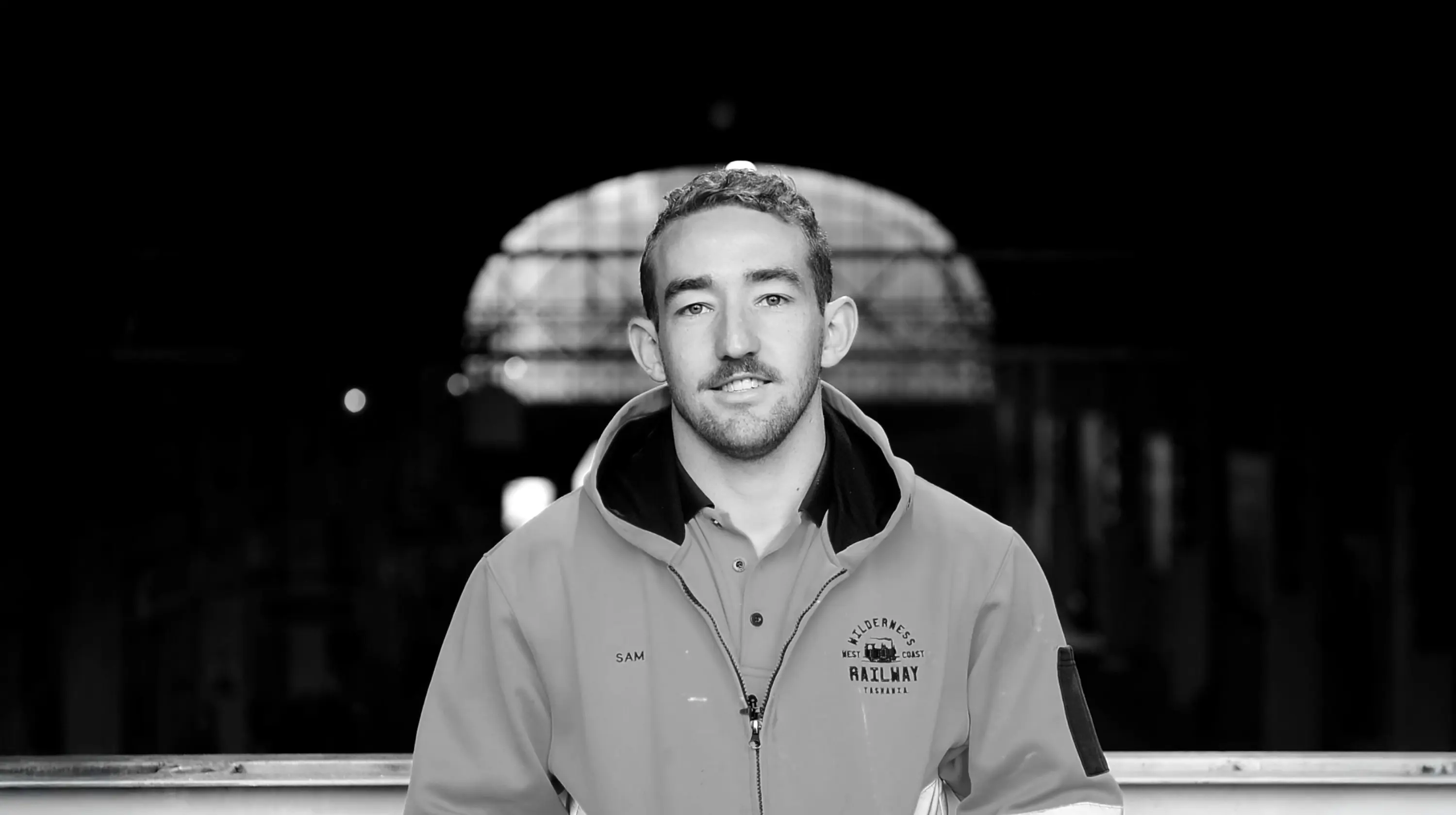 A man wearing a jacket with the West Coast Wilderness Railway and his name, 'SAM', embroidered on it. Behind him is a dark railway station.