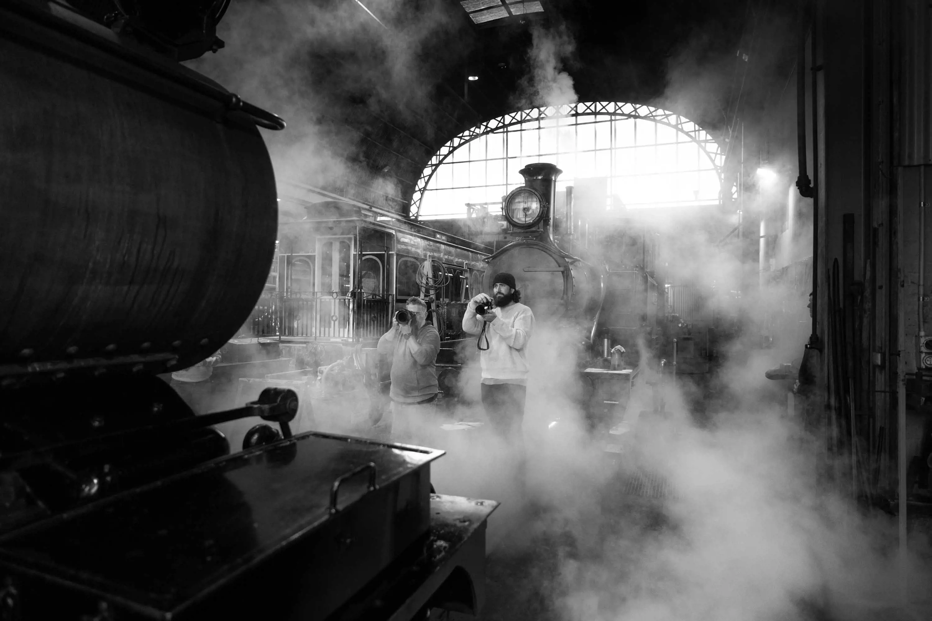 Two photographers take photos while they stand in the steam from a locomotive under the covered roof of an old station.