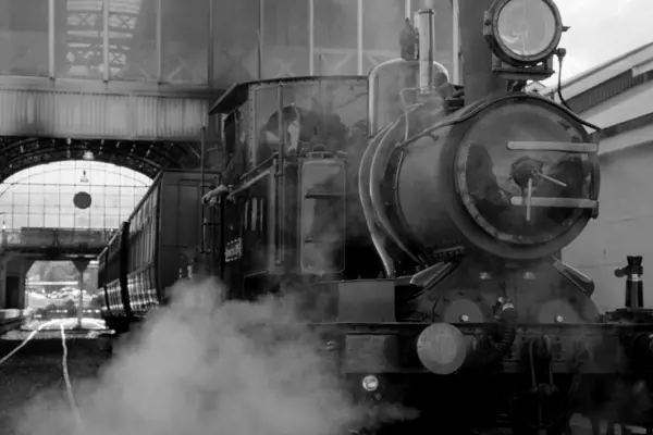A large steam locomotive with passenger carriages exits the station with a puff of steam.