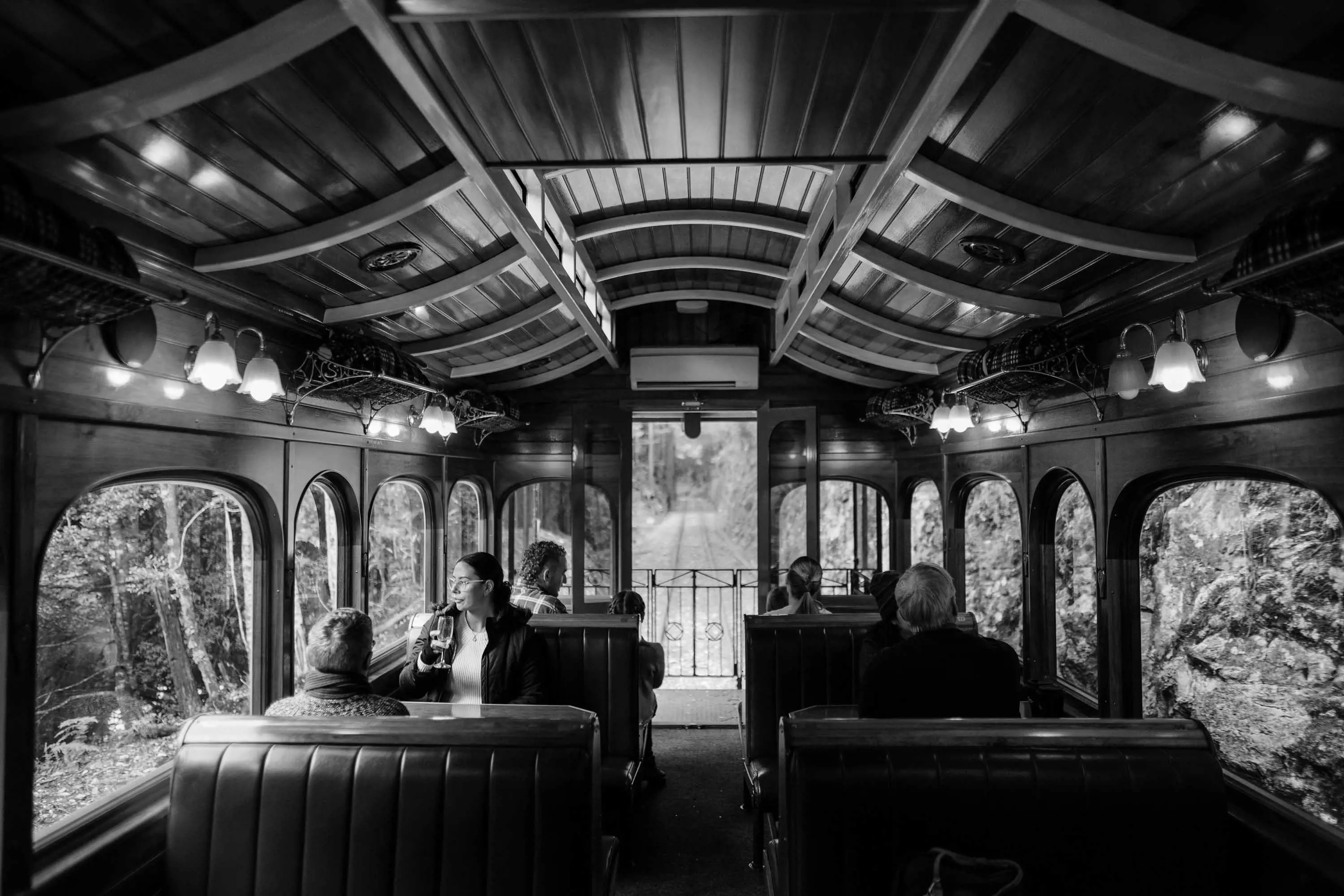 The wooden interior of a restored passenger carriage with large, arched windows looking out at dense rainforest. 