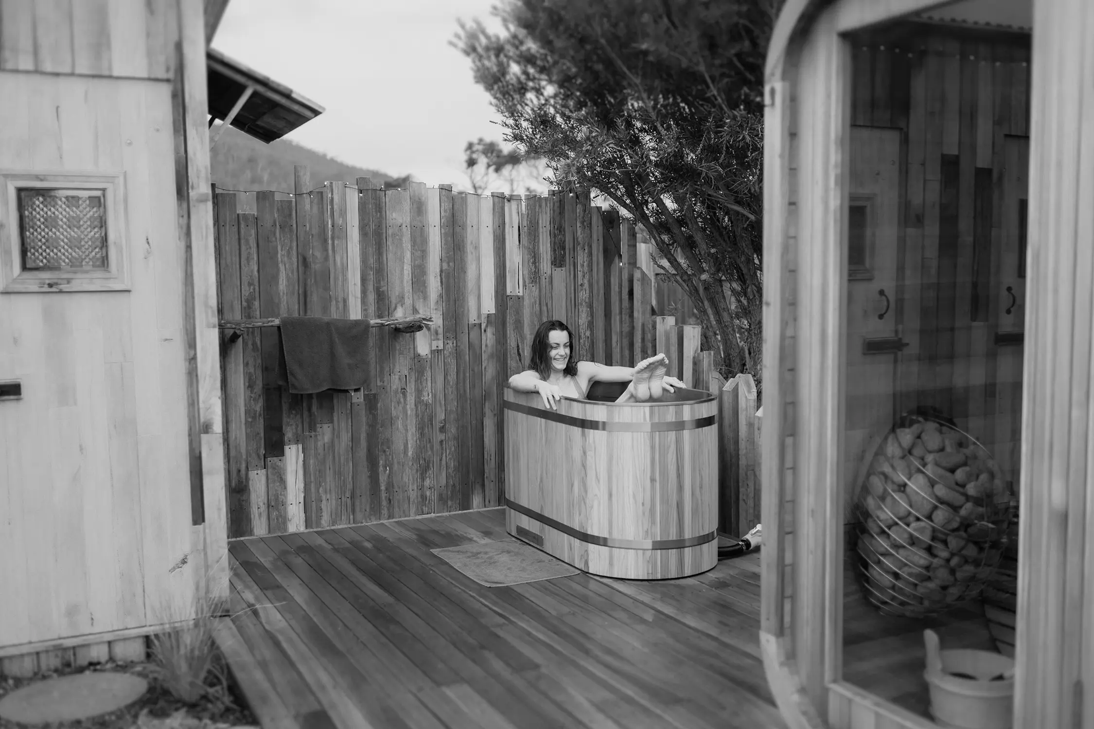 On an outdoor deck in a secluded backyard, a woman relaxes in a wood-clad outdoor bathtub.