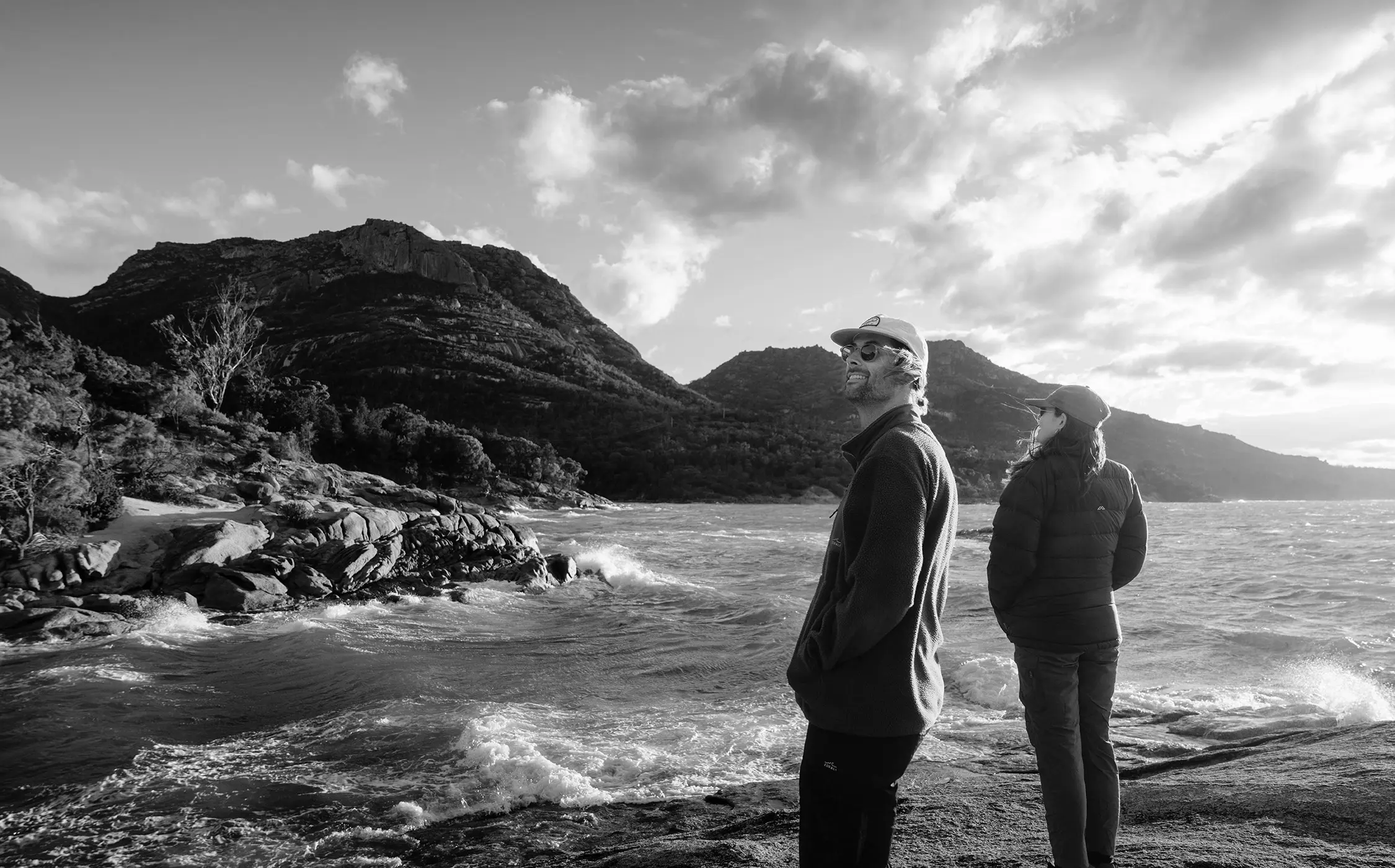 A man and a woman stand on rocks at a beach, watching the waves crashing onto the shore below them. Mountainous terrain rises beyond.