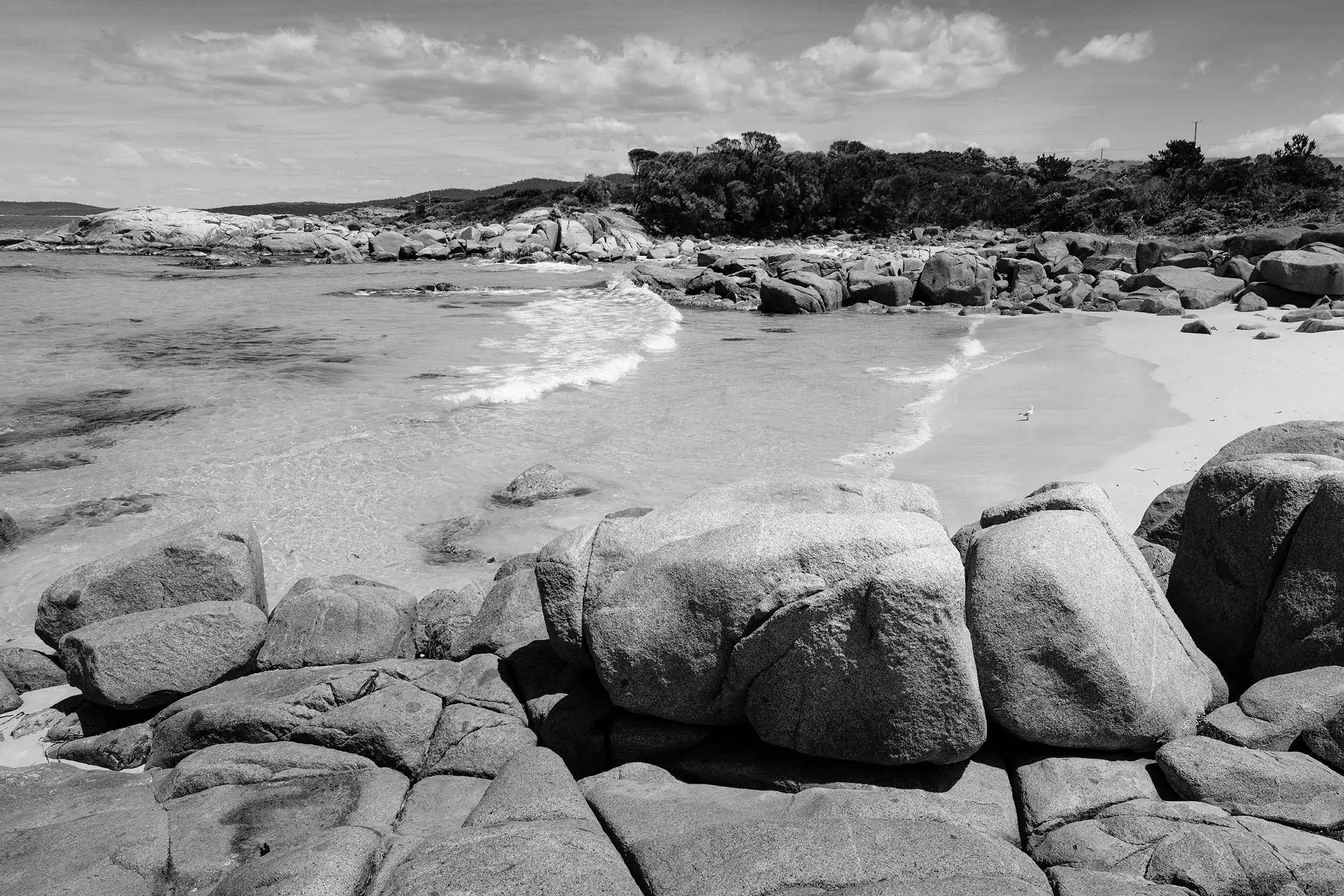 A calm, clear beach scattered with piles of angular rocks.