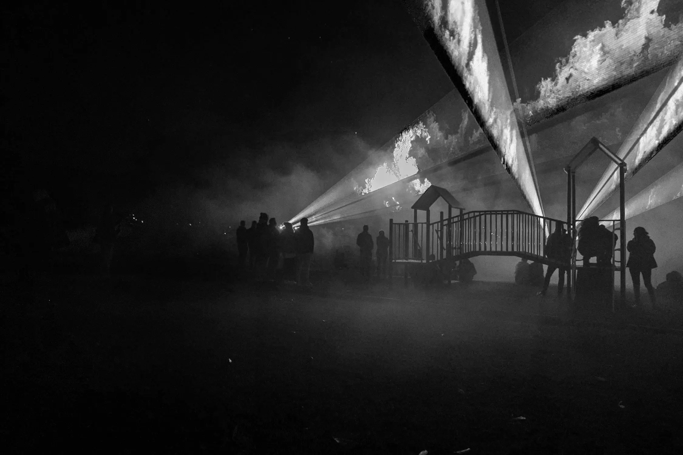 In a dark, smoke-filled playground, lasers shine into the night sky, images of flames projected onto them.