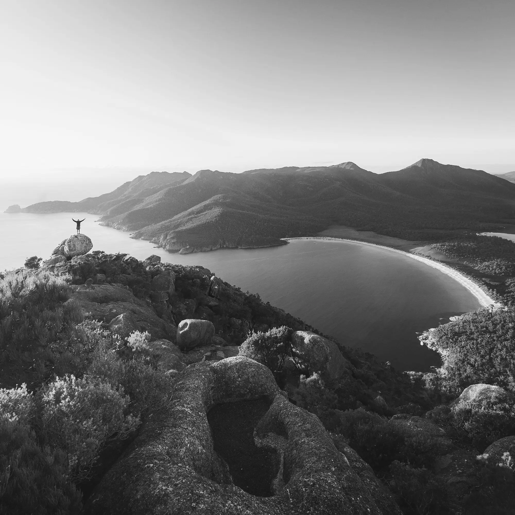 A person stands atop a boulder on a huge cliff, dropping down below to reveal a beautiful ocean bay with a mountainous background.