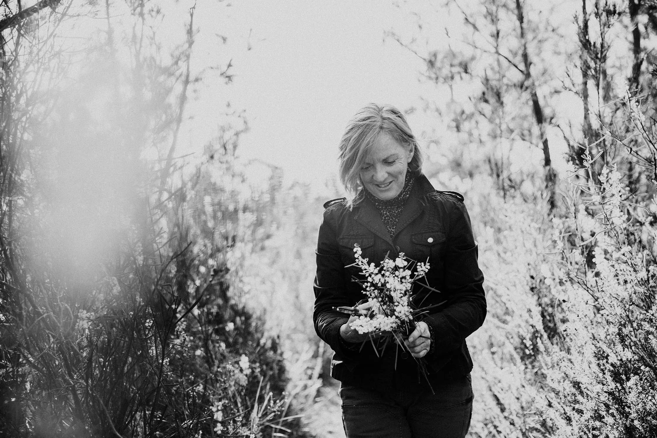 A woman in a jacket walks through trees, the sun shining through the branches onto the bundle of florals she is holding.