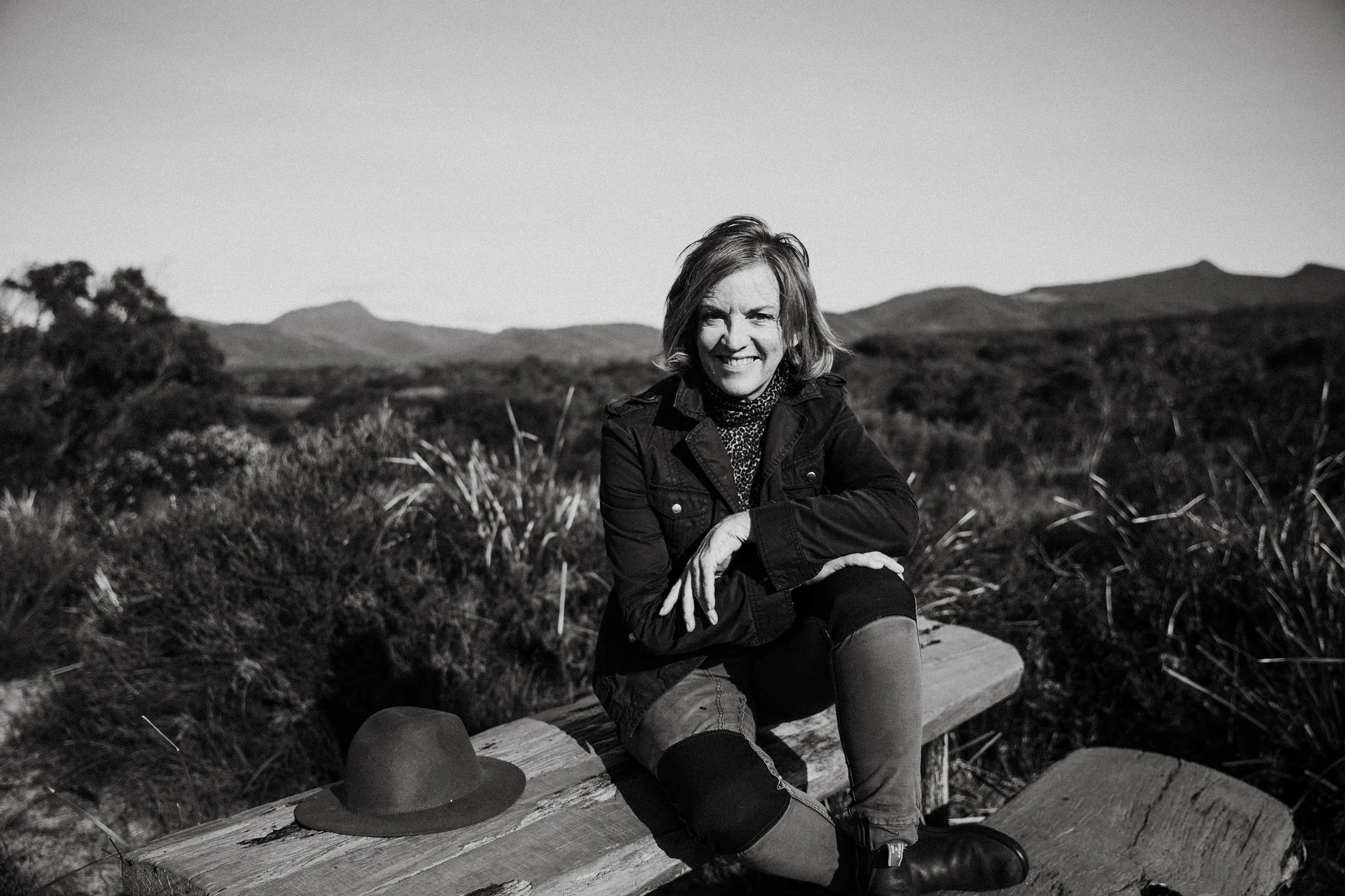A woman sits on a picnic table, her legs on the bench, and her hat resting on the table beside her. The environment is beachside scrub with mountains on the far horizon.