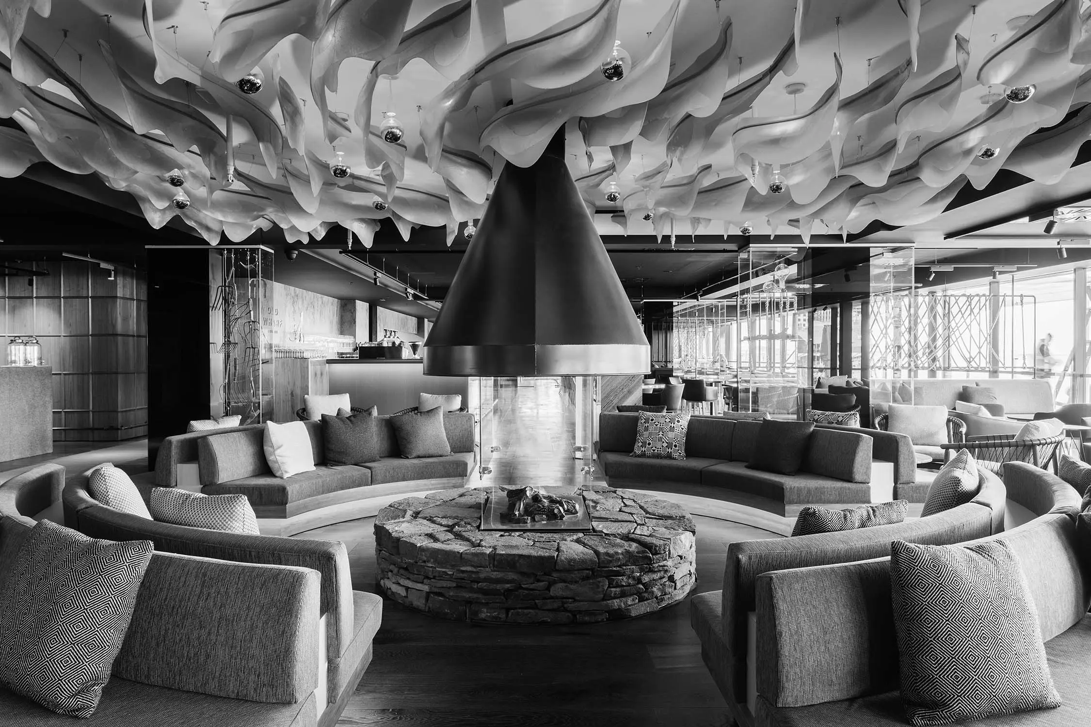 A striking circular fireplace inside a hotel lobby. Long cosy couches circle around the stone plinth, with a huge rangehood hanging overhead, disappearing into wavy sculptures hung from the ceiling.