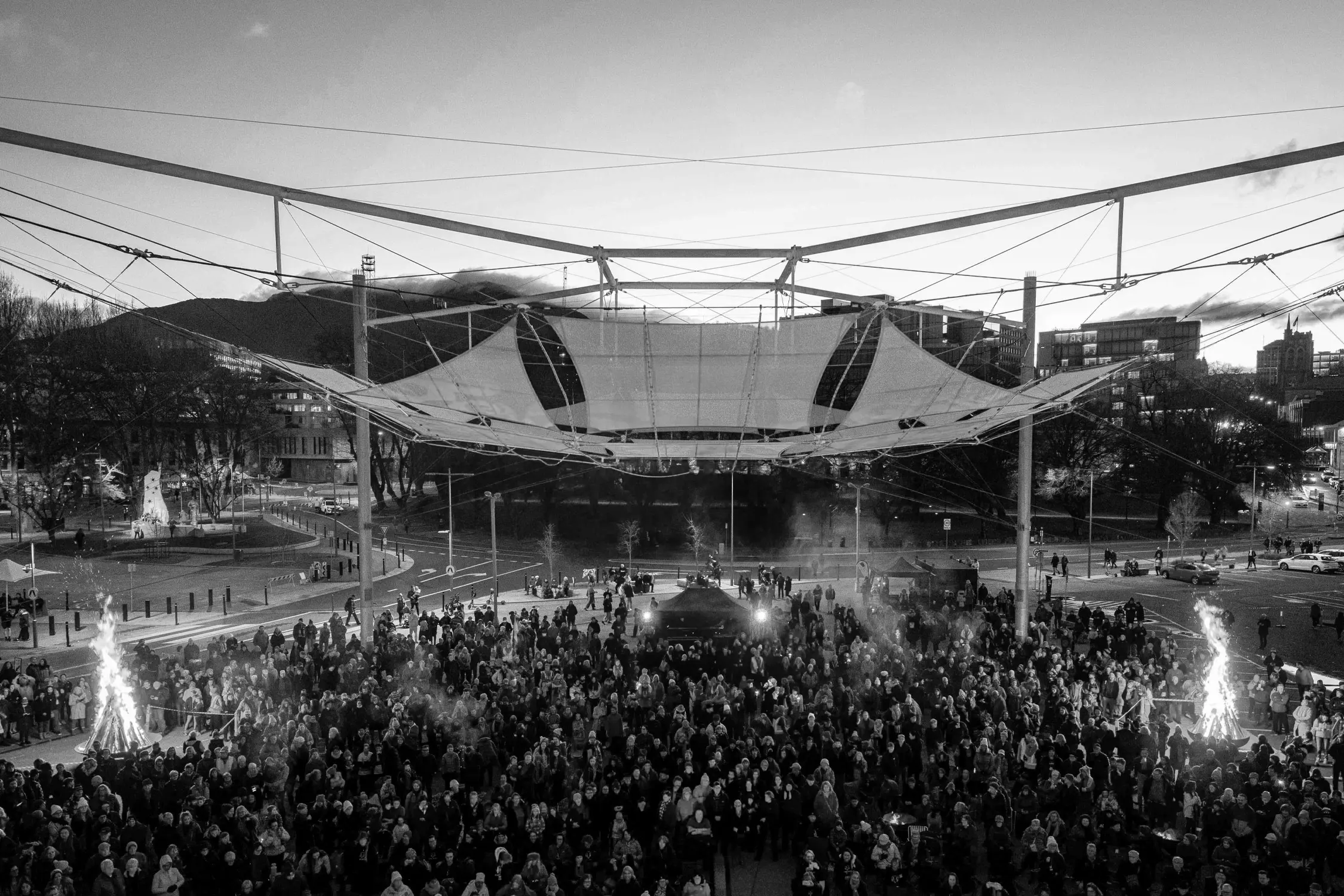A large outdoor crowd gathered at dusk for a festival under a glowing canopy structure. Twp bonfires at each end radiate warmth, and the twilight sky enhances the festive atmosphere.