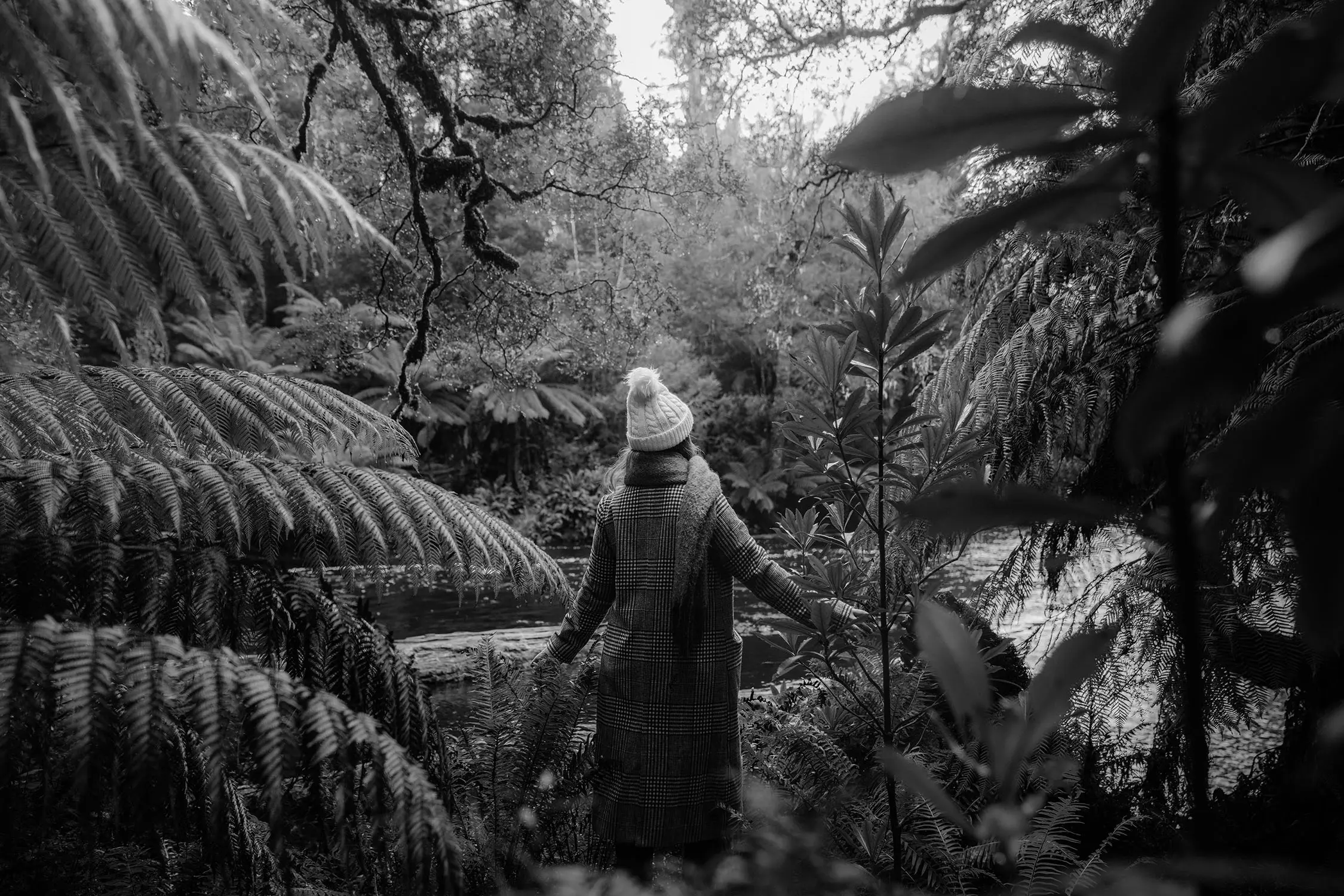 A woman in a beanie and scarf walks alongside a river, surrounded by ferns and low-hanging trees glistening with moisture.