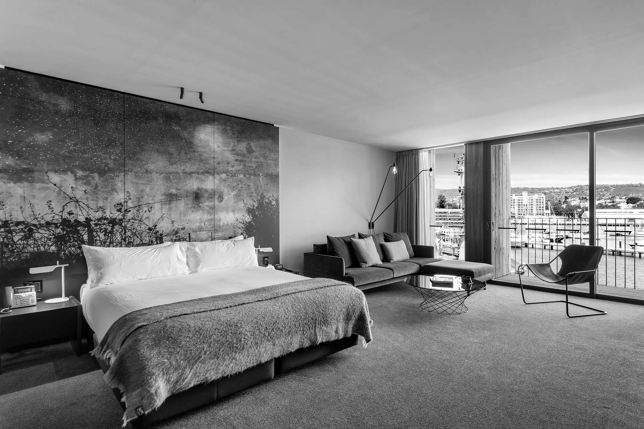 The interior of a spacious hotel room, with stylish furniture and floor-to-ceiling windows looking out over a harbour.