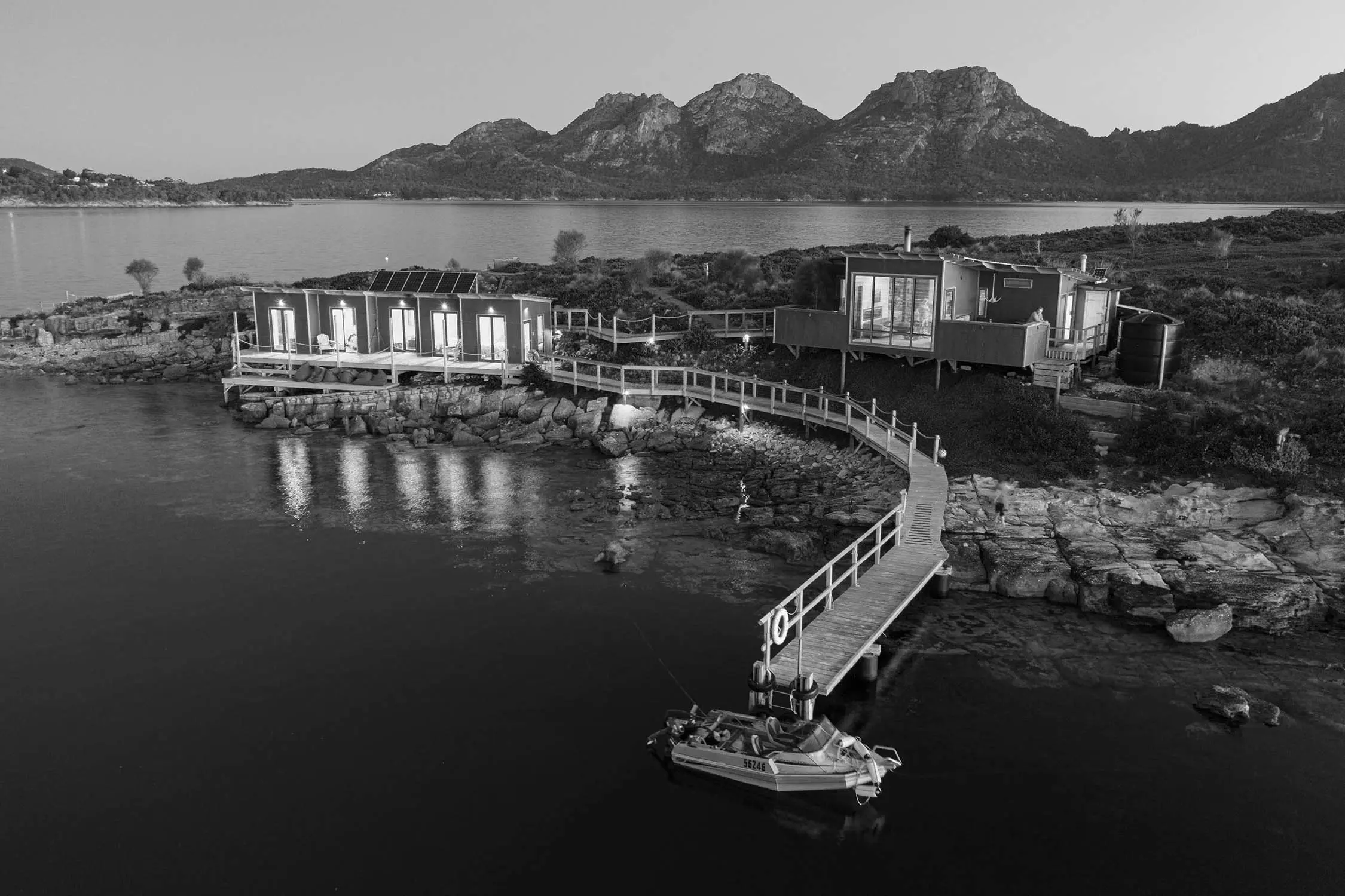 An aerial black and white view shows modern eco-lodges connected by wooden boardwalks on a rocky Tasmanian peninsula. Mountains rise across the calm waters while a small boat rests at the jetty.