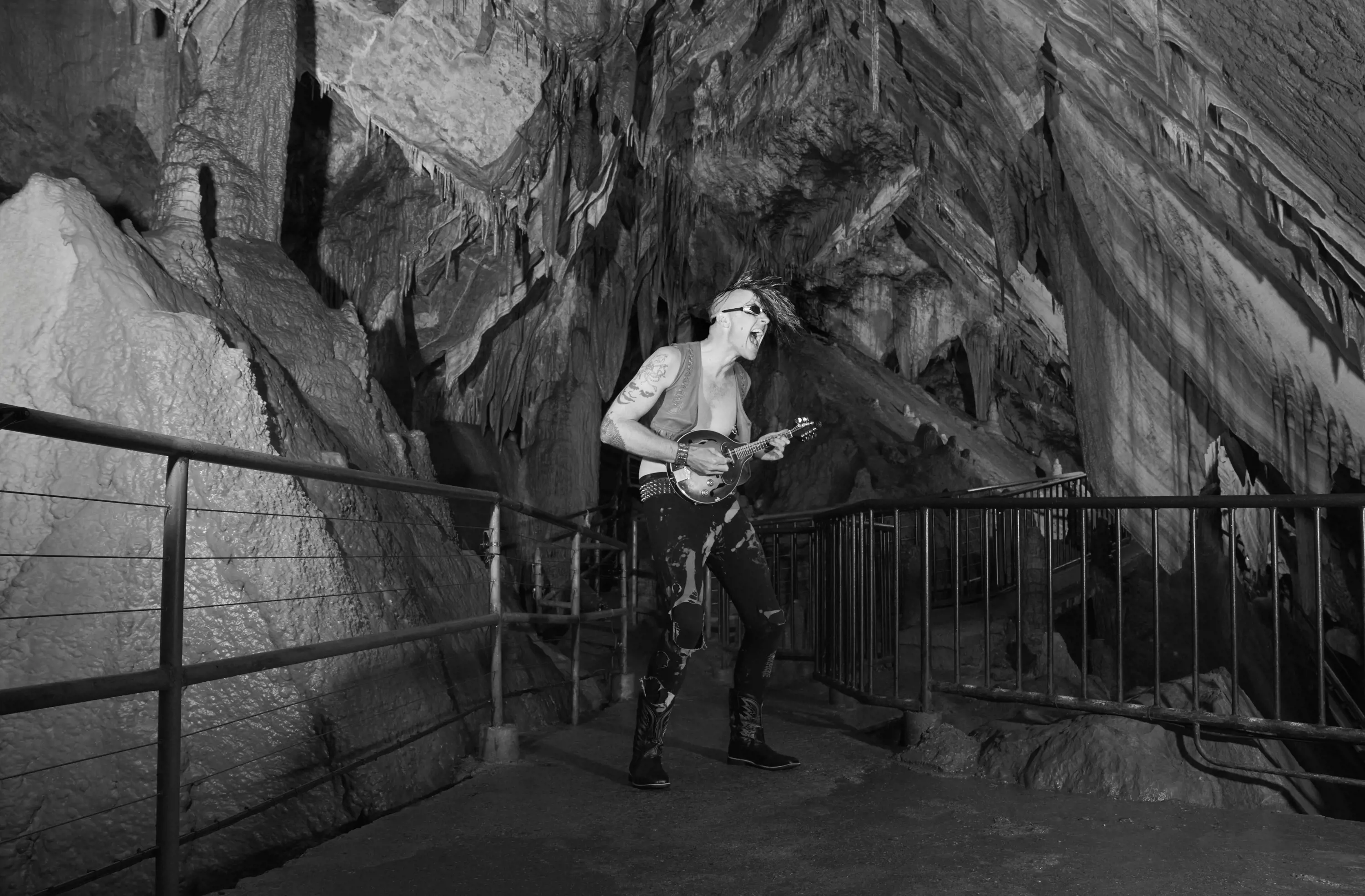 A man with tattoos and a mohawk wearing a vest and torn jeans, plays a banjo and sings on a walkway inside an enormous cave.  