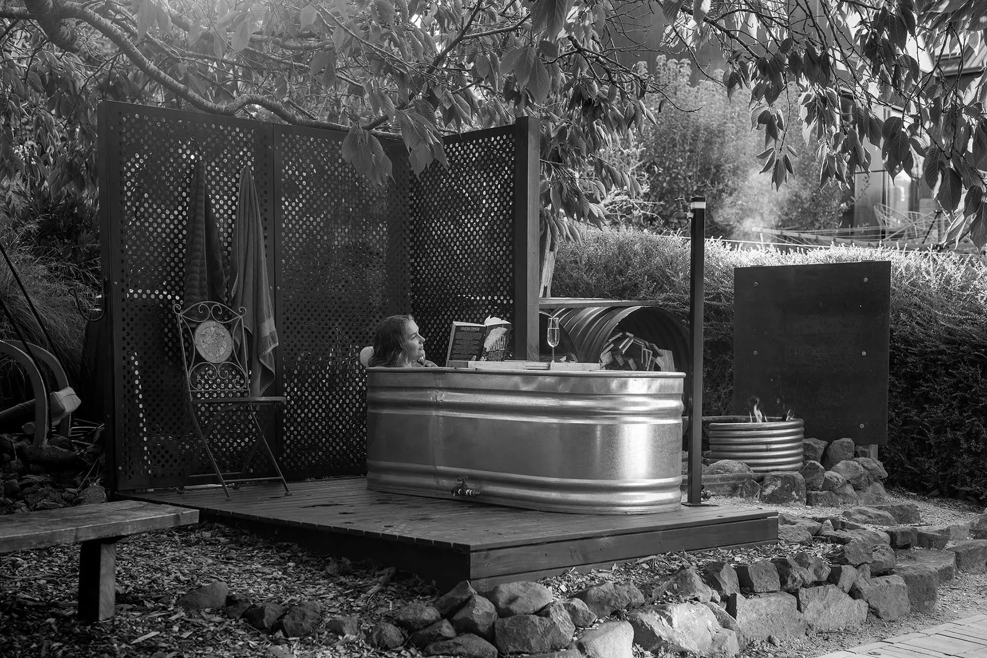A person relaxes in a metal stock tank bath on a wooden deck, surrounded by lattice privacy screens, native vegetation, and a small water feature.