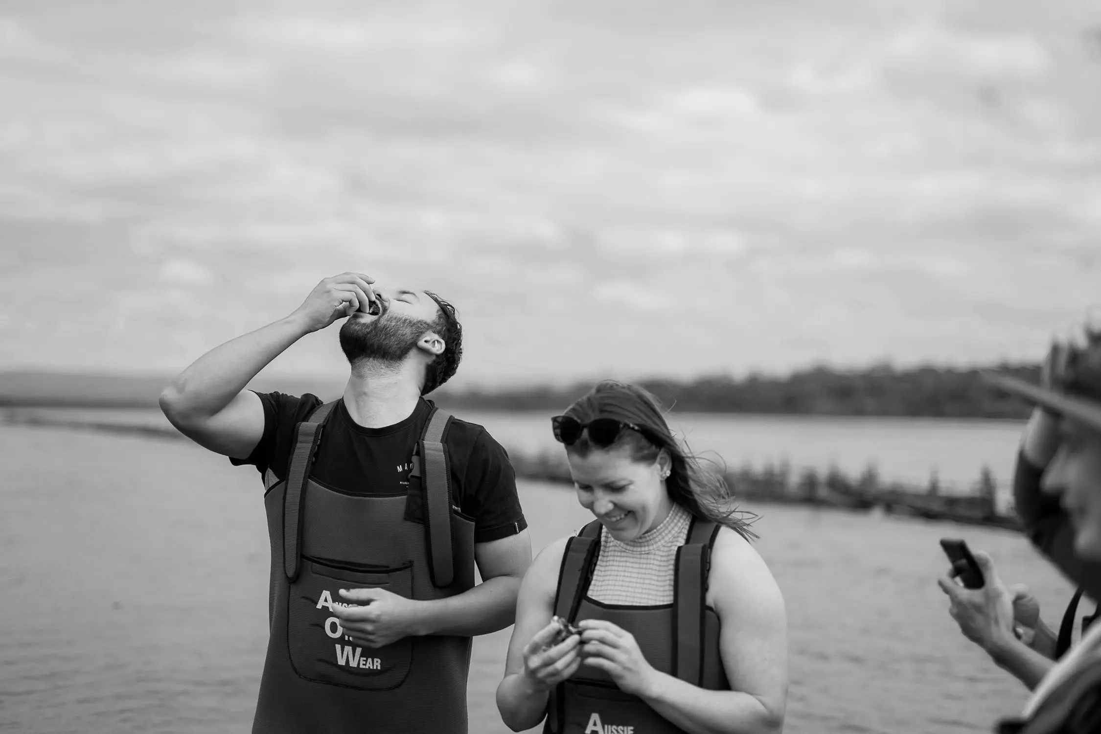 Two people wearing wading overalls are slurping oysters straight from their shells. In the background are the still waters of an ocean's inlet.