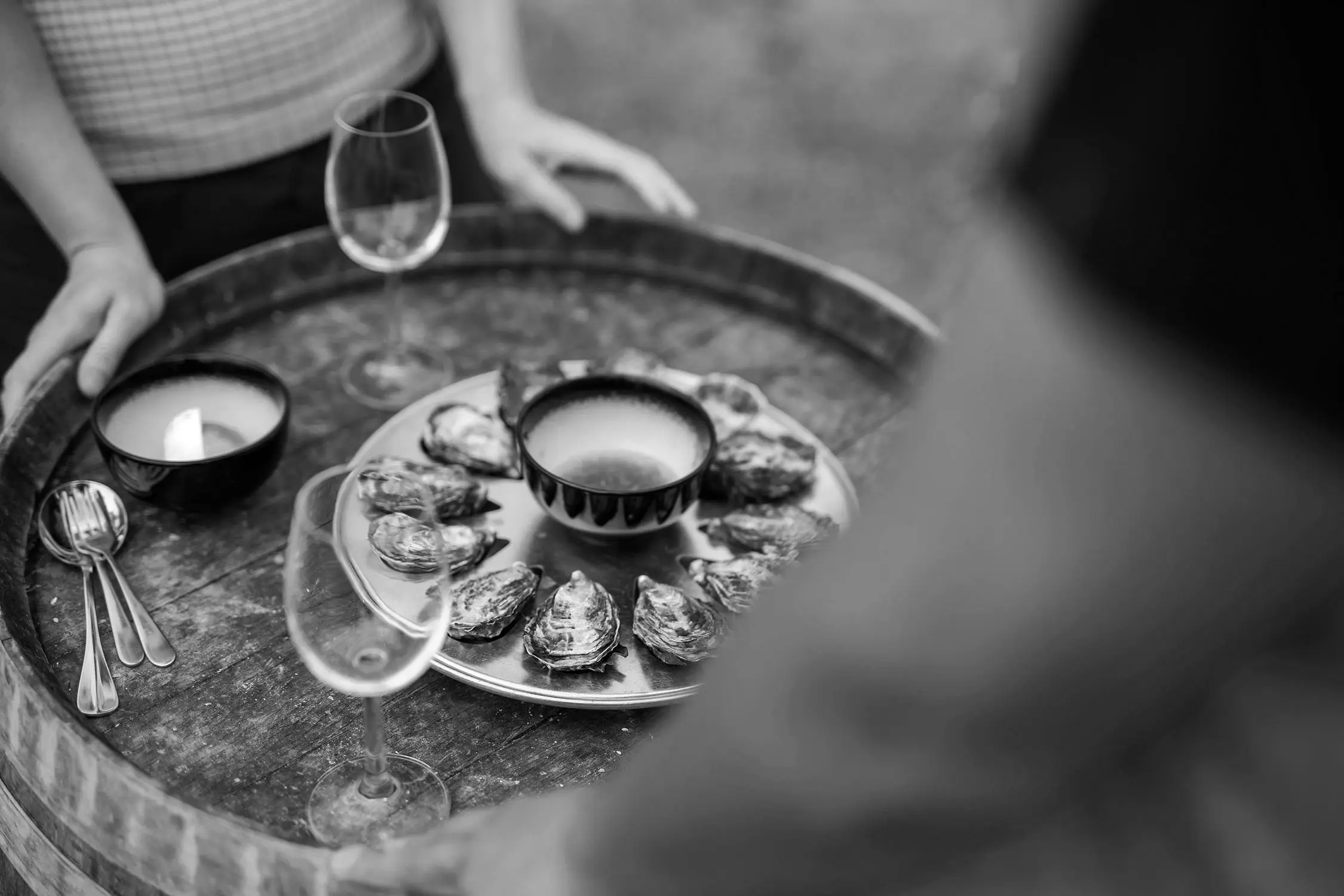 A black and white overhead view shows a wooden barrel topped with fresh oysters arranged on a round plate. Wine glasses and cutlery are placed on the weathered surface.
