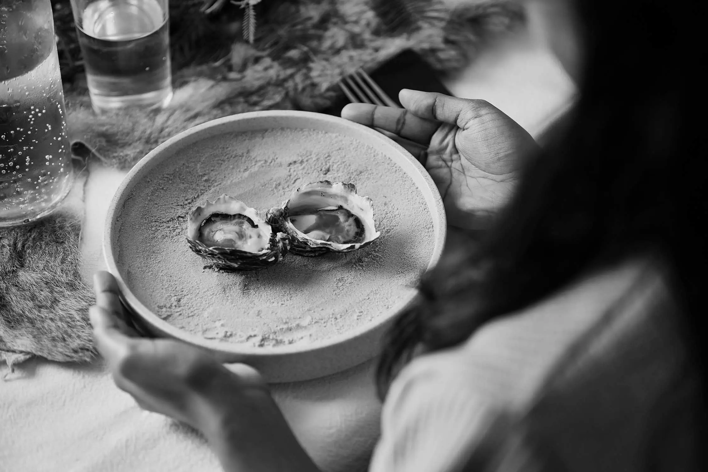A person holds a textured ceramic plate with two shucked oysters sitting in their shells. On the table, sparkling water in a bottle and an animal pelt are visible.
