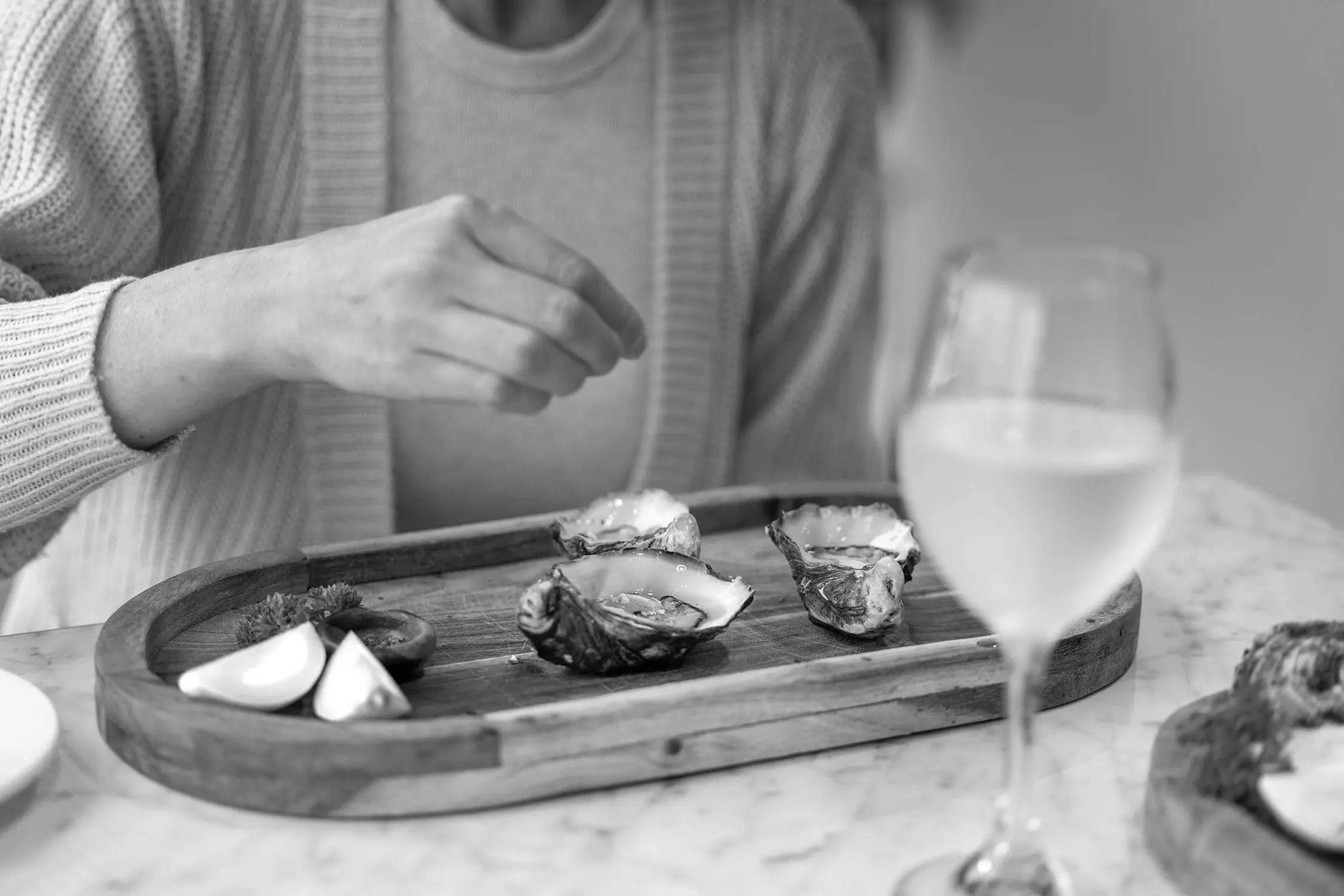 A black and white photograph shows a person reaching toward fresh oysters on a wooden serving board. Lemon wedges accompany the oysters, with wine glasses visible in the background.
